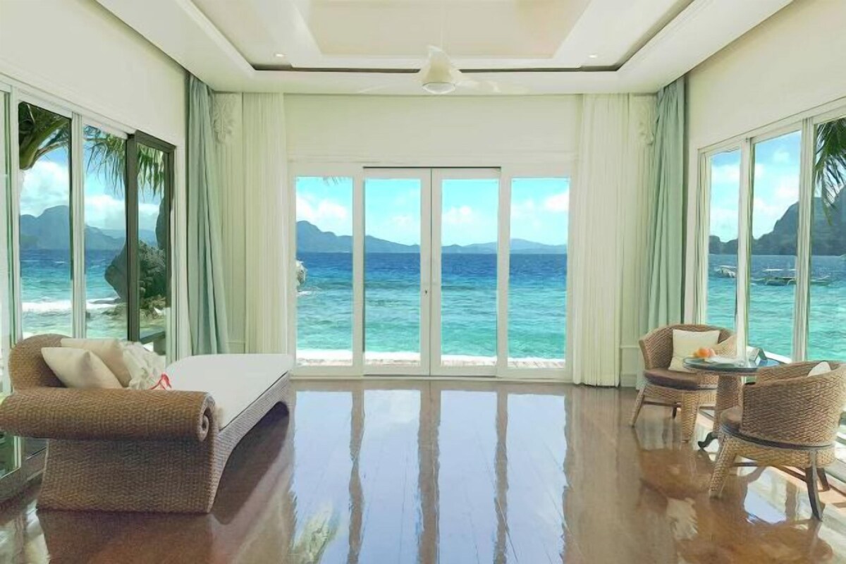 A room with a large sliding glass door leading to a balcony with a view of the ocean and mountains. There is a bed, a chair, and a small table with a vase of flowers.