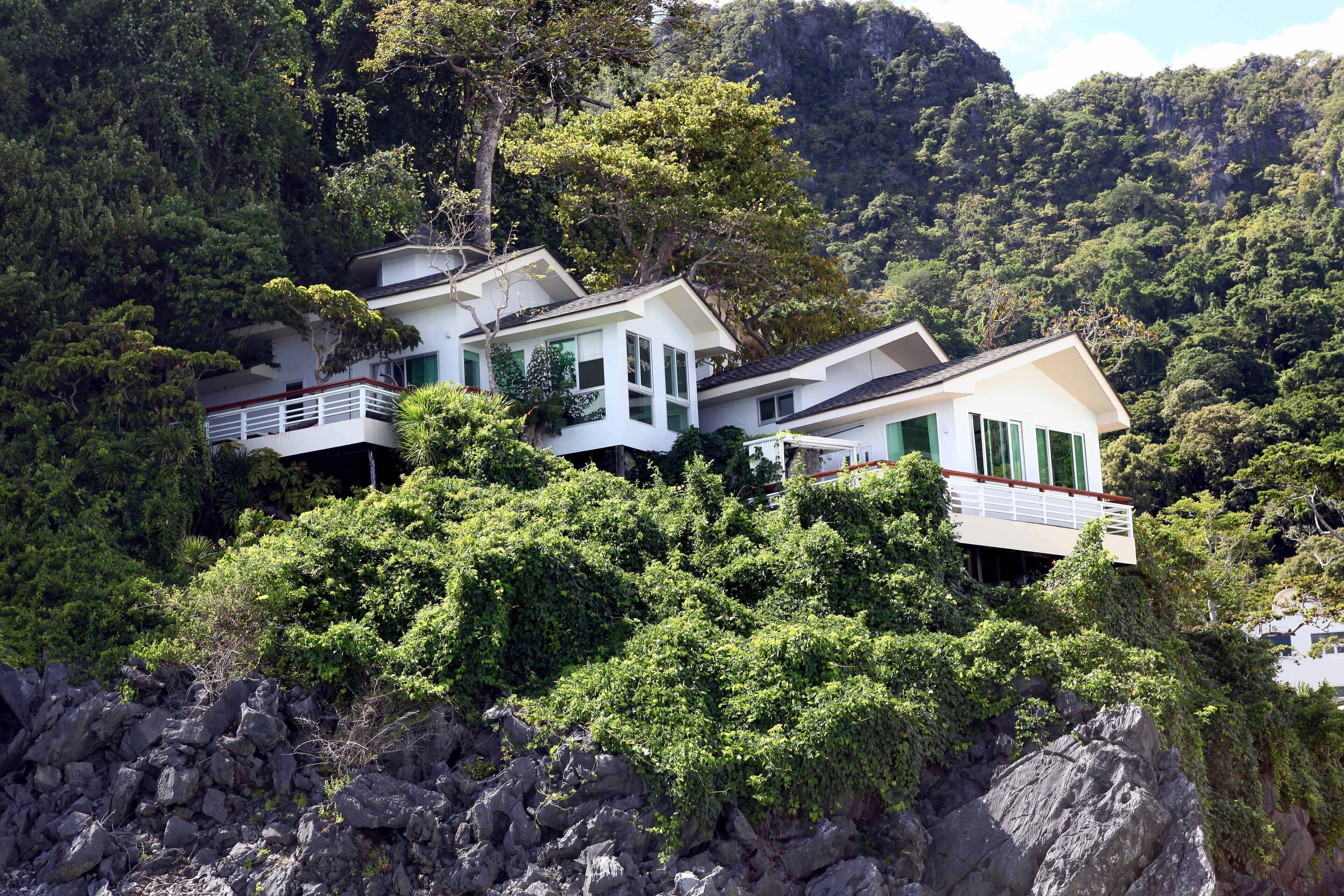A multi-story house with a balcony, surrounded by lush greenery and rocky terrain.