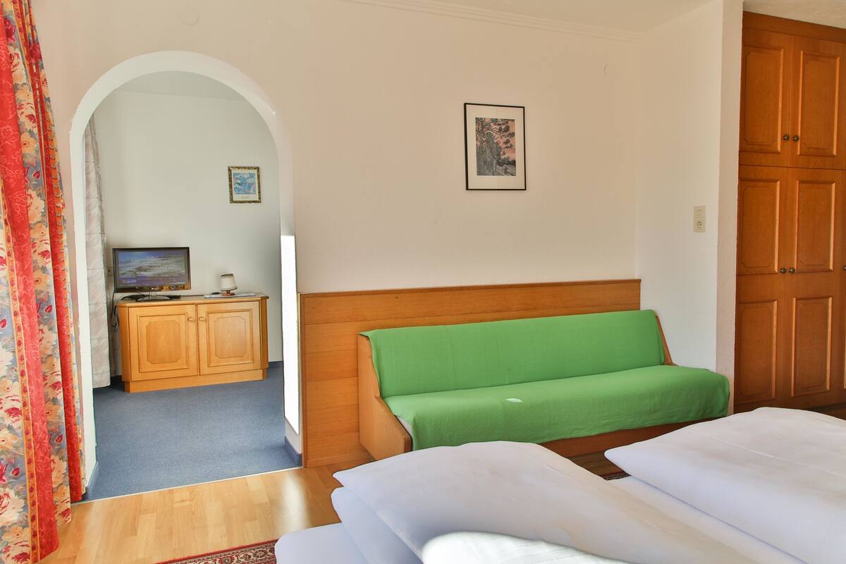 Habitación de hotel con un armario de madera, un sofá verde, una cama con ropa blanca y un televisor sobre un mueble de madera.