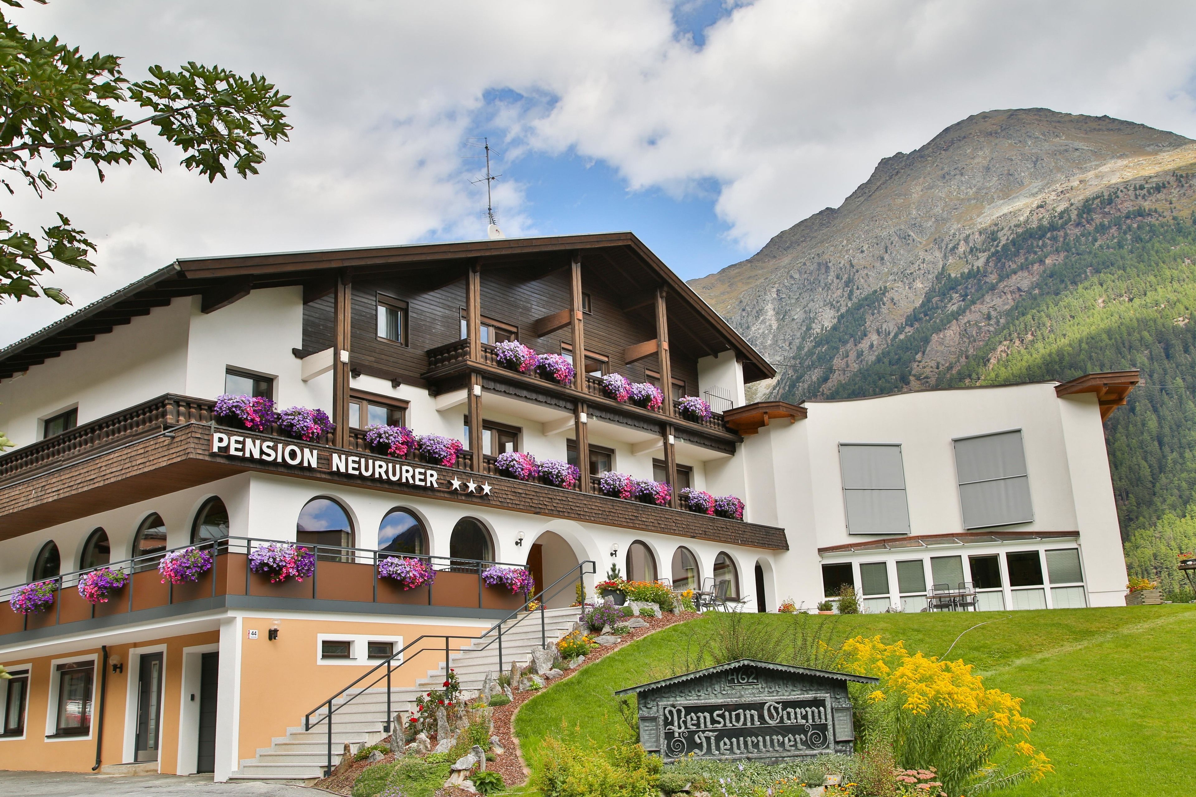 A two-story building with balconies adorned with flowers, a sign reading "Pension Neururer," and a mountainous backdrop.