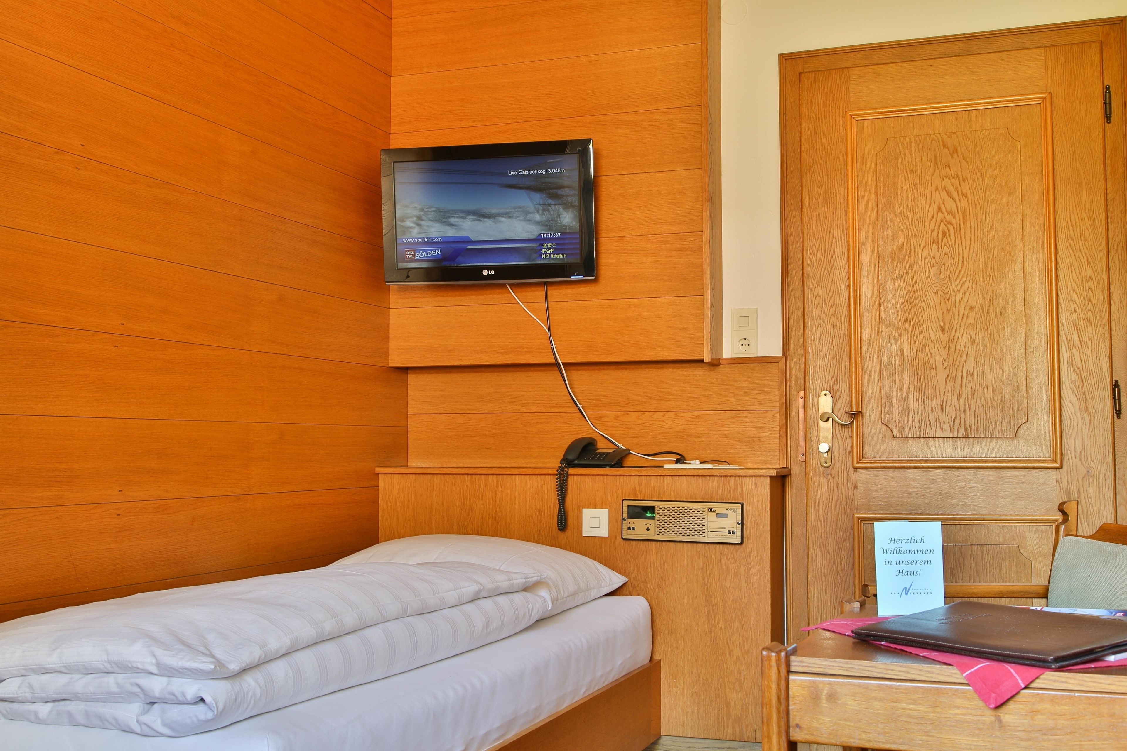 A hotel room with a bed, a desk with a laptop, a television mounted on the wall, and a wooden door.