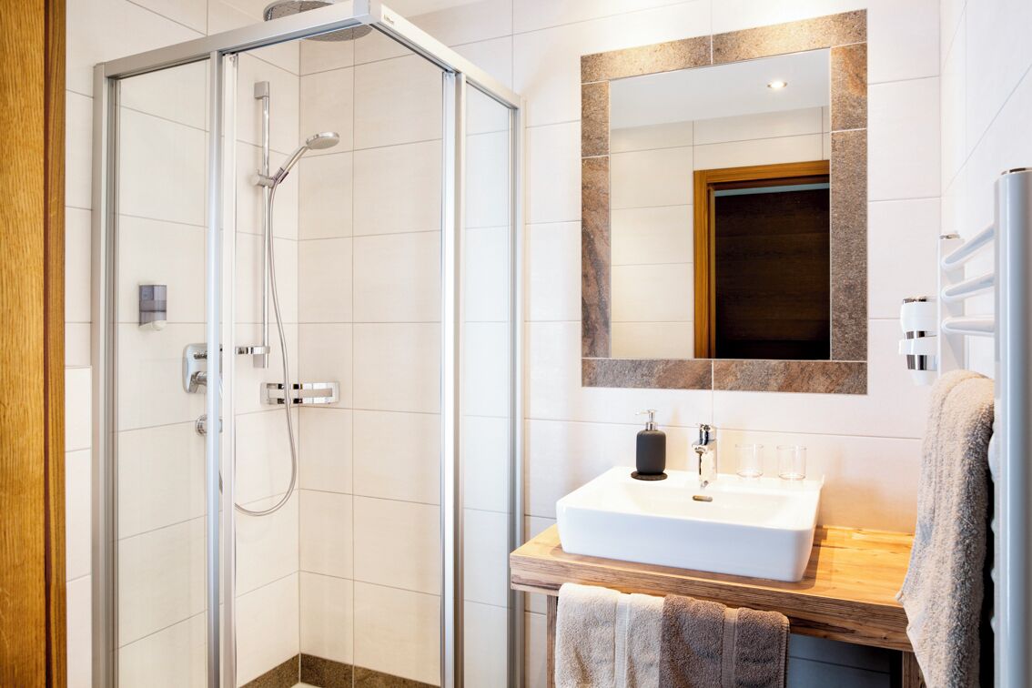 A modern bathroom with a glass-enclosed shower, a wooden vanity with a white sink, and a large mirror with a stone frame.