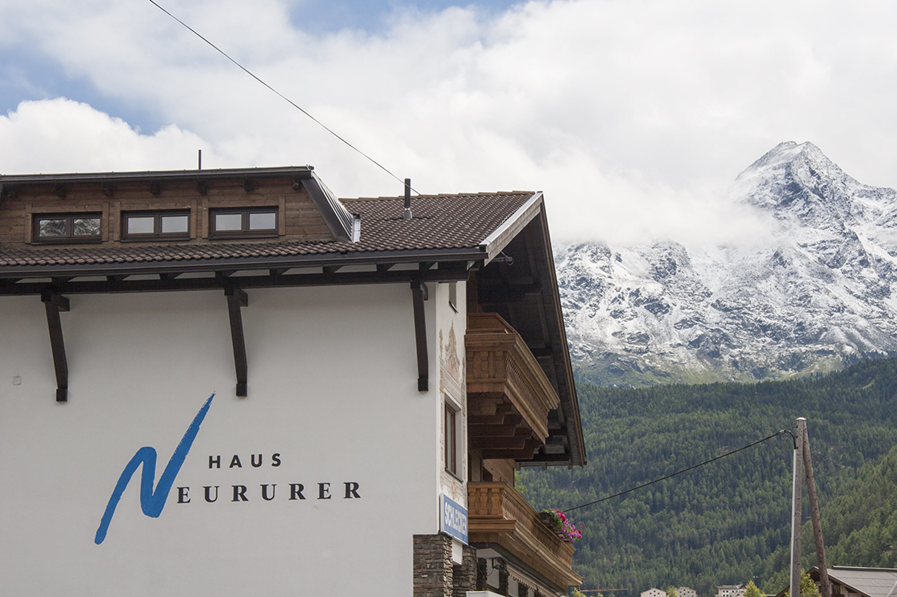 A building with a sign reading "HAUS NEURUER" and a view of snow-capped mountains in the background.