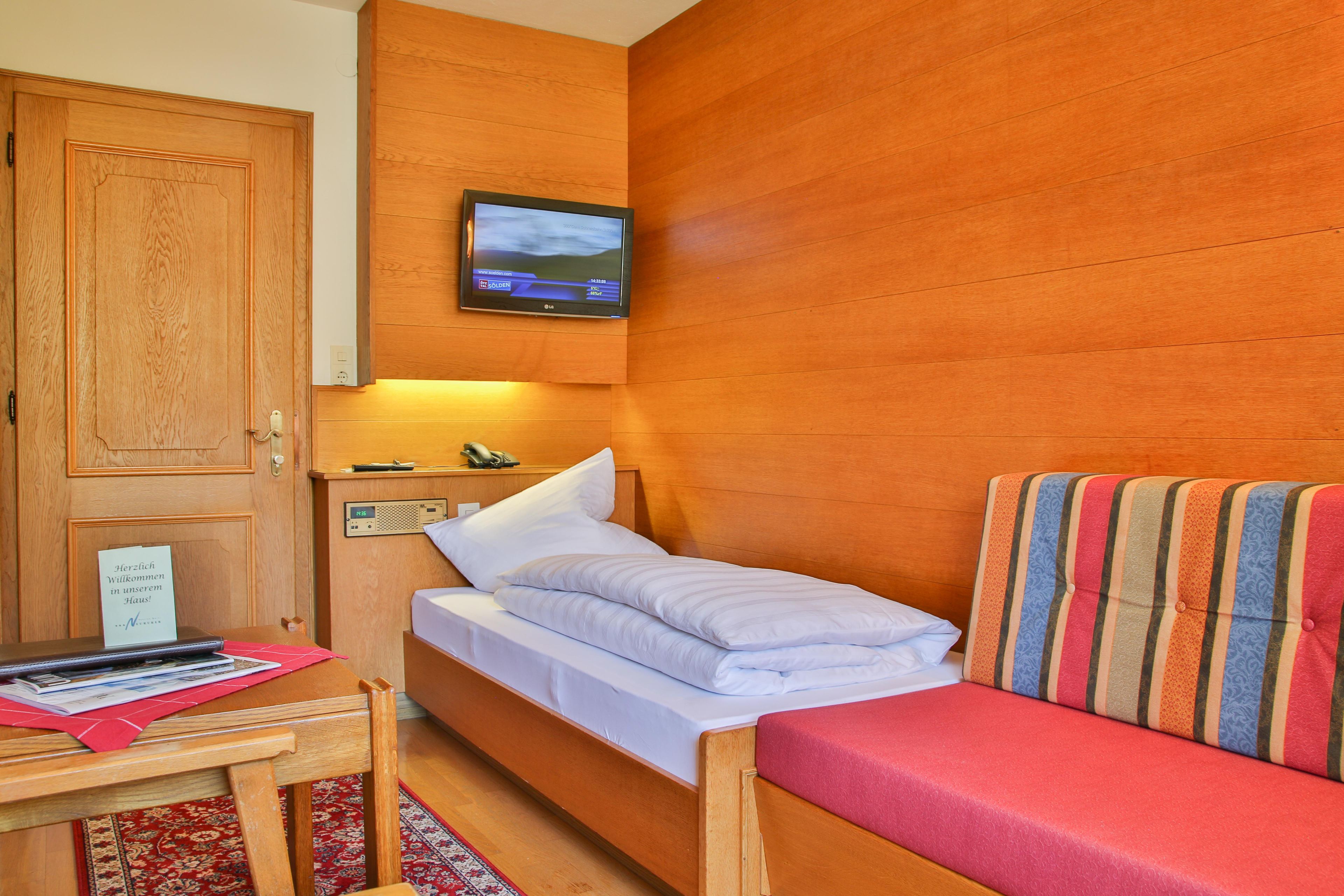 Una habitación con pared de madera, una cama con ropa de cama blanca, un escritorio pequeño con una computadora portátil, un televisor montado sobre la cama y un cojín a rayas rojas en un banco de madera.