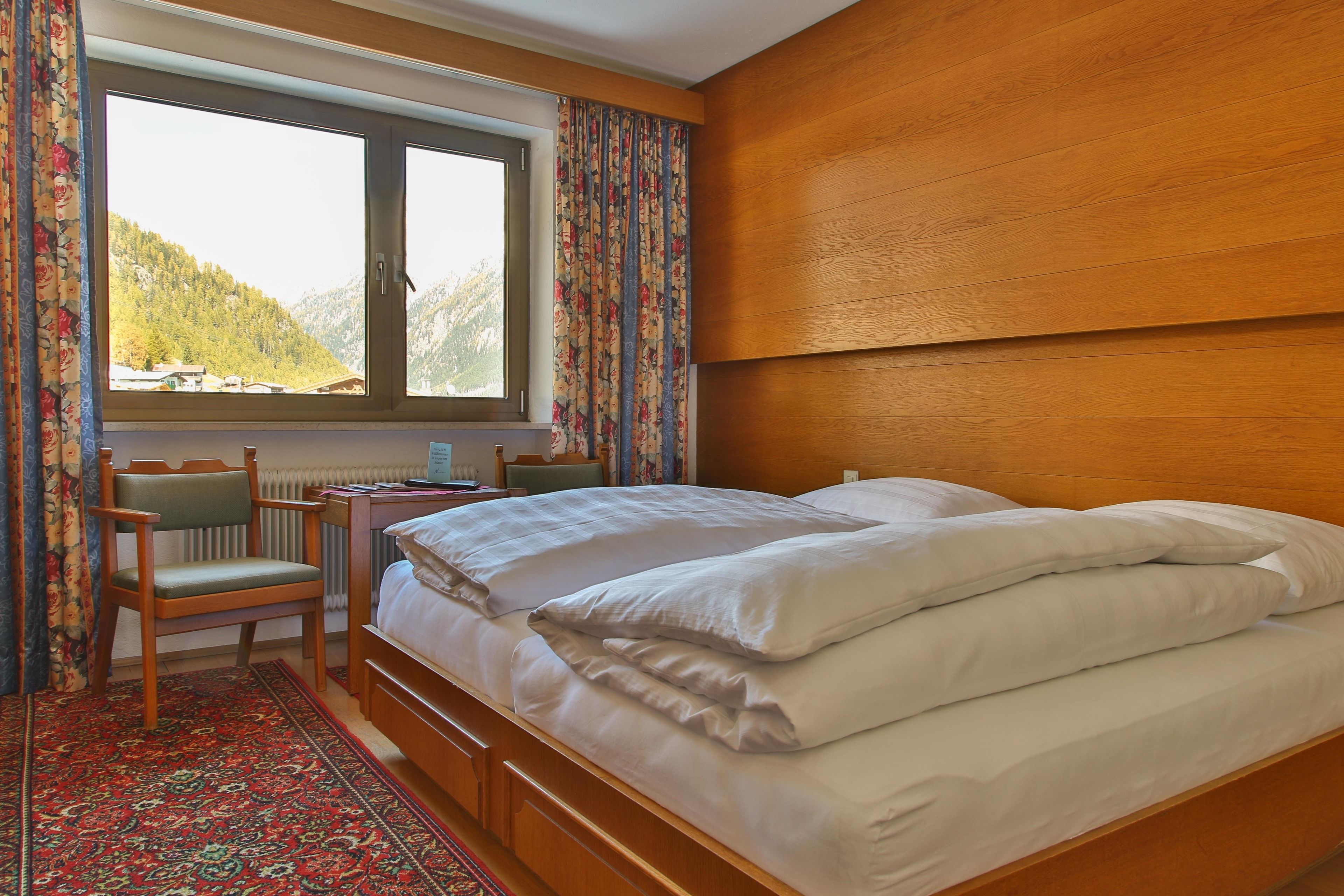 A bedroom with a wooden headboard, a bed with white bedding, a desk with a chair, and a window with curtains.