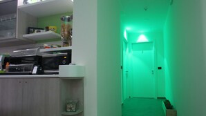 Eine Küche mit Mikrowelle, Kaffeemaschine und diversen Küchenutensilien in Regalen. Ein Flur mit einem grünen Licht, das zu einer Tür führt.