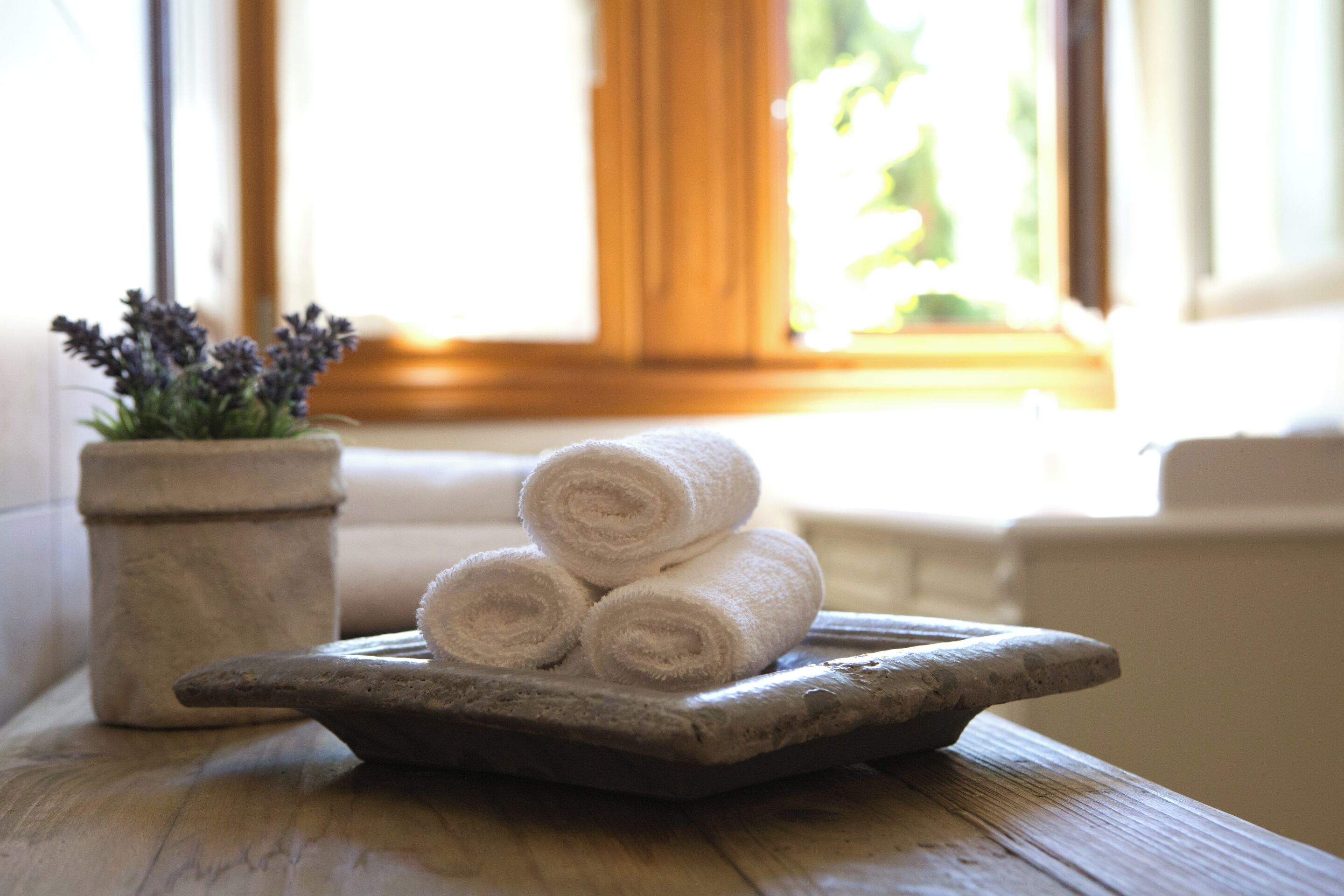 A wooden tray with rolled white towels on a wooden table near a window with a view of greenery.