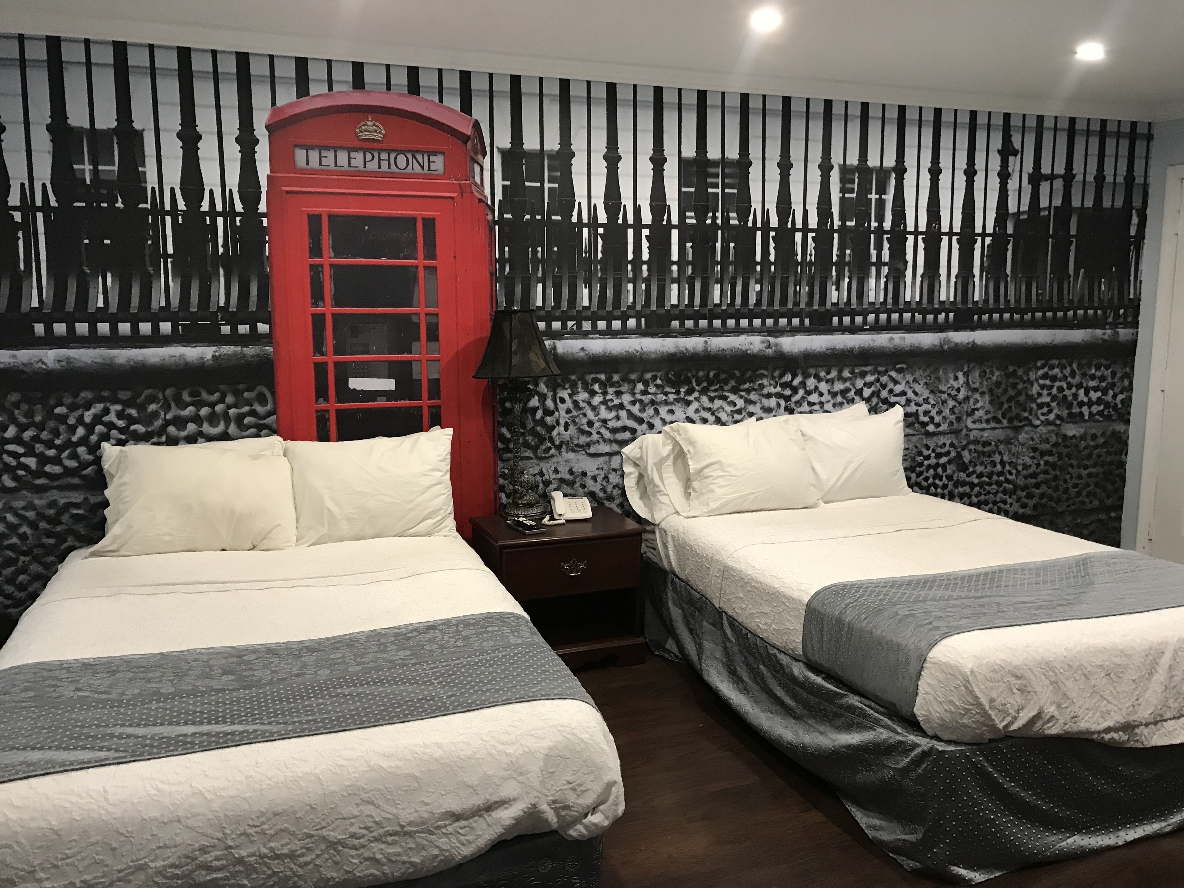 A hotel room with two beds, a red telephone booth, a patterned wall, and a nightstand with a lamp.