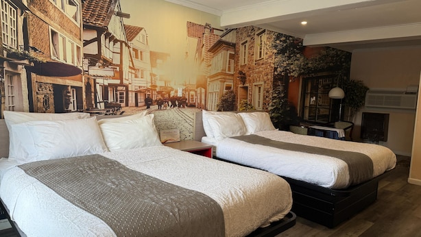 Una habitación de hotel con dos camas, un mural de una escena callejera y una mesita con una lámpara.