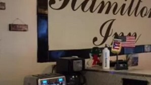 Una habitación con un letrero grande que dice "Hamilton Inn" y una pequeña bandera estadounidense. Hay una máquina de café y un cuadro en el mostrador.