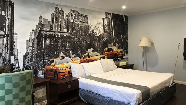 A hotel room with a large cityscape mural, a bed with a grey and white bedspread, a wooden nightstand, a green checkered chair, and a floor lamp.