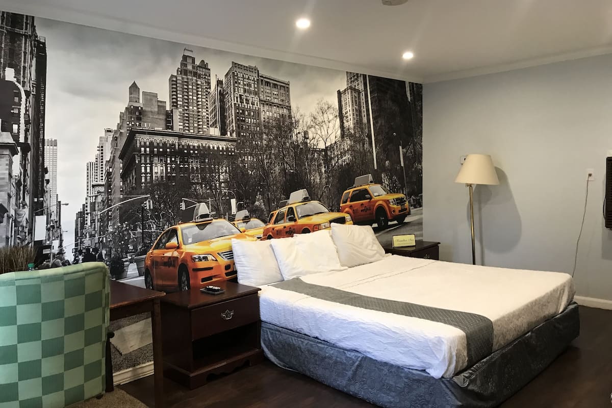 A hotel room with a large cityscape mural, a bed with a grey and white bedspread, a wooden nightstand, a green checkered chair, and a floor lamp.