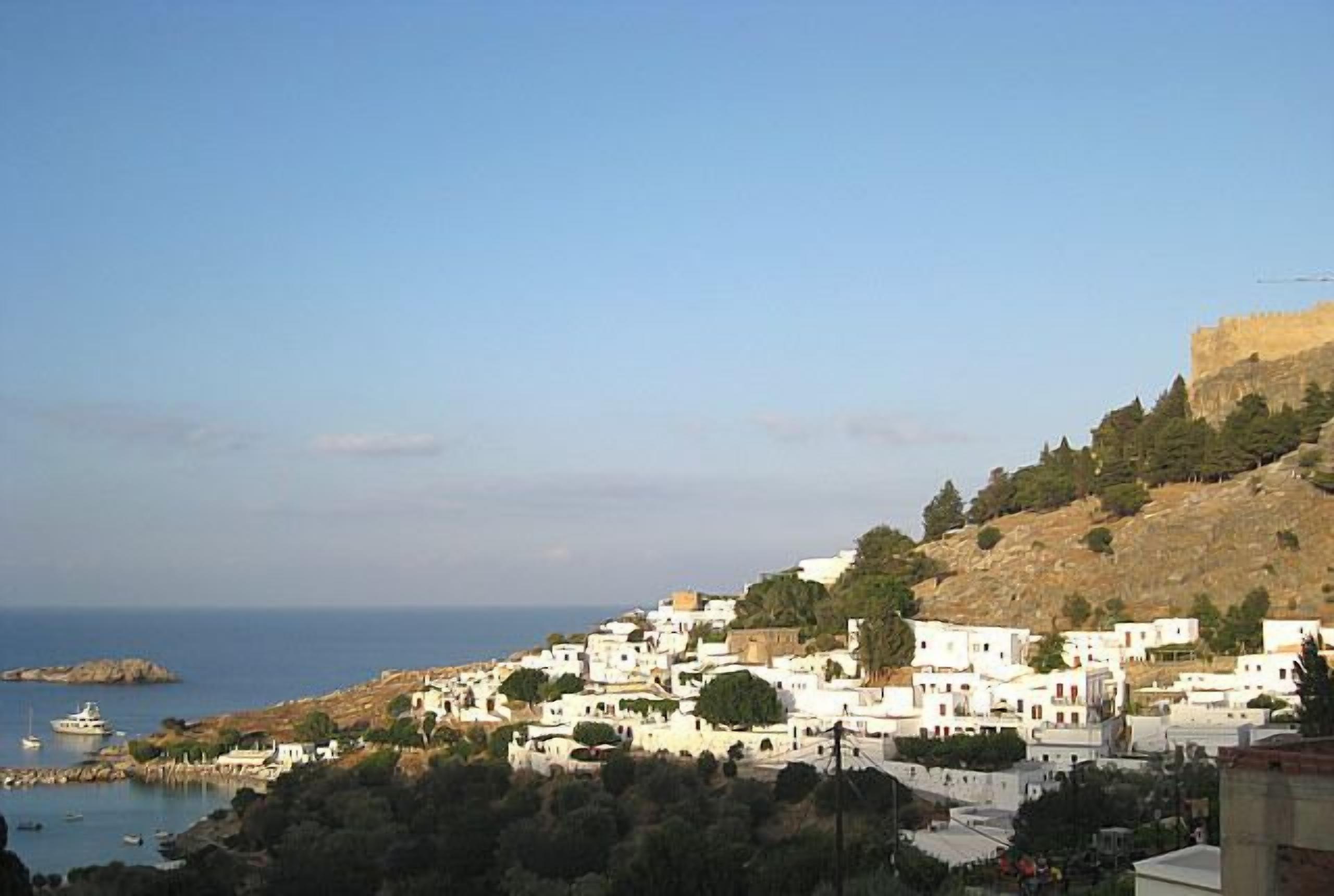 A coastal town with white buildings, a rocky hill, and a clear sky.