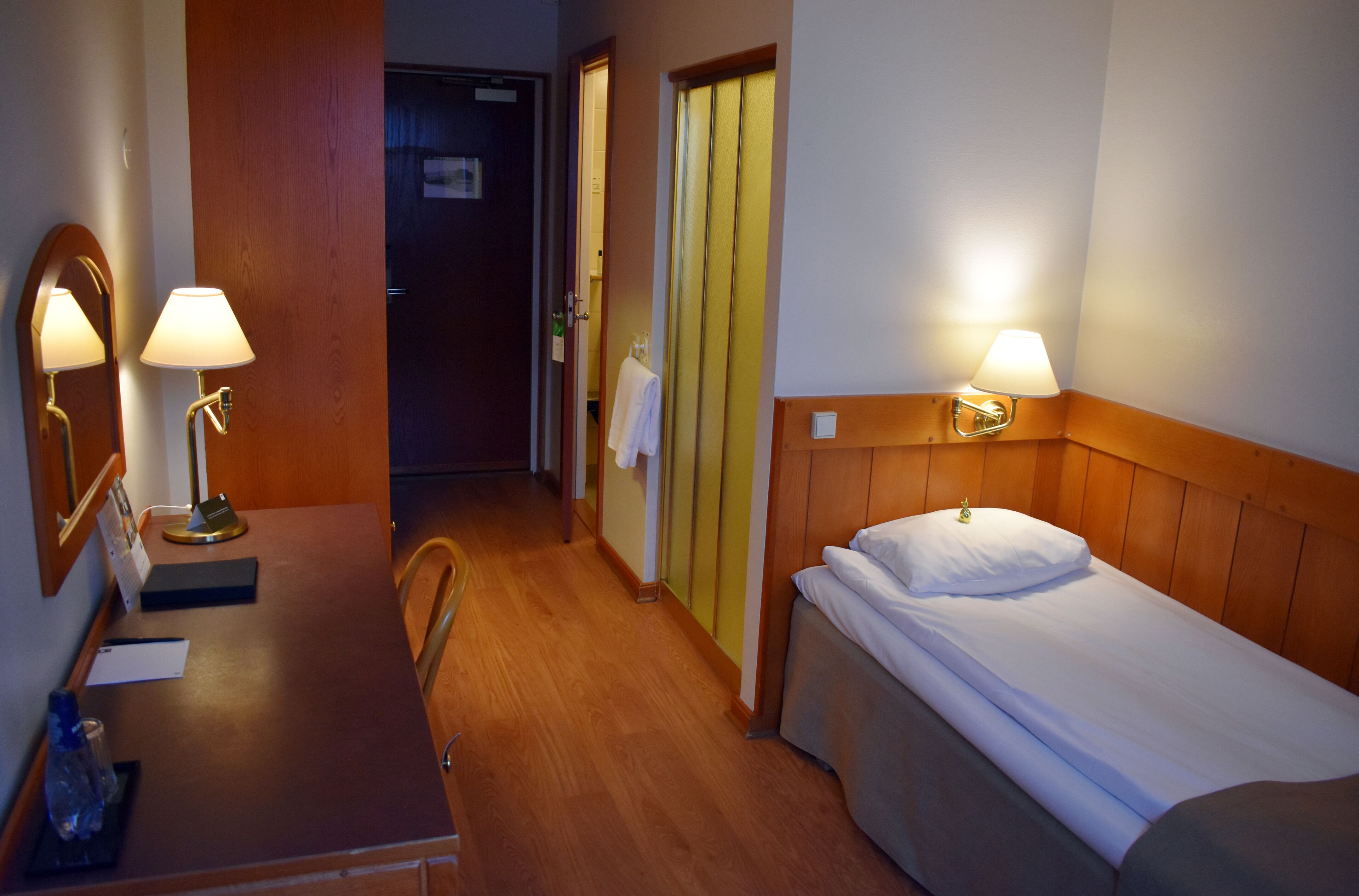 A hotel room with a wooden floor, a bed with white bedding, two bedside lamps, a wooden headboard, a wooden desk with a mirror, a chair, and a door leading to another room.