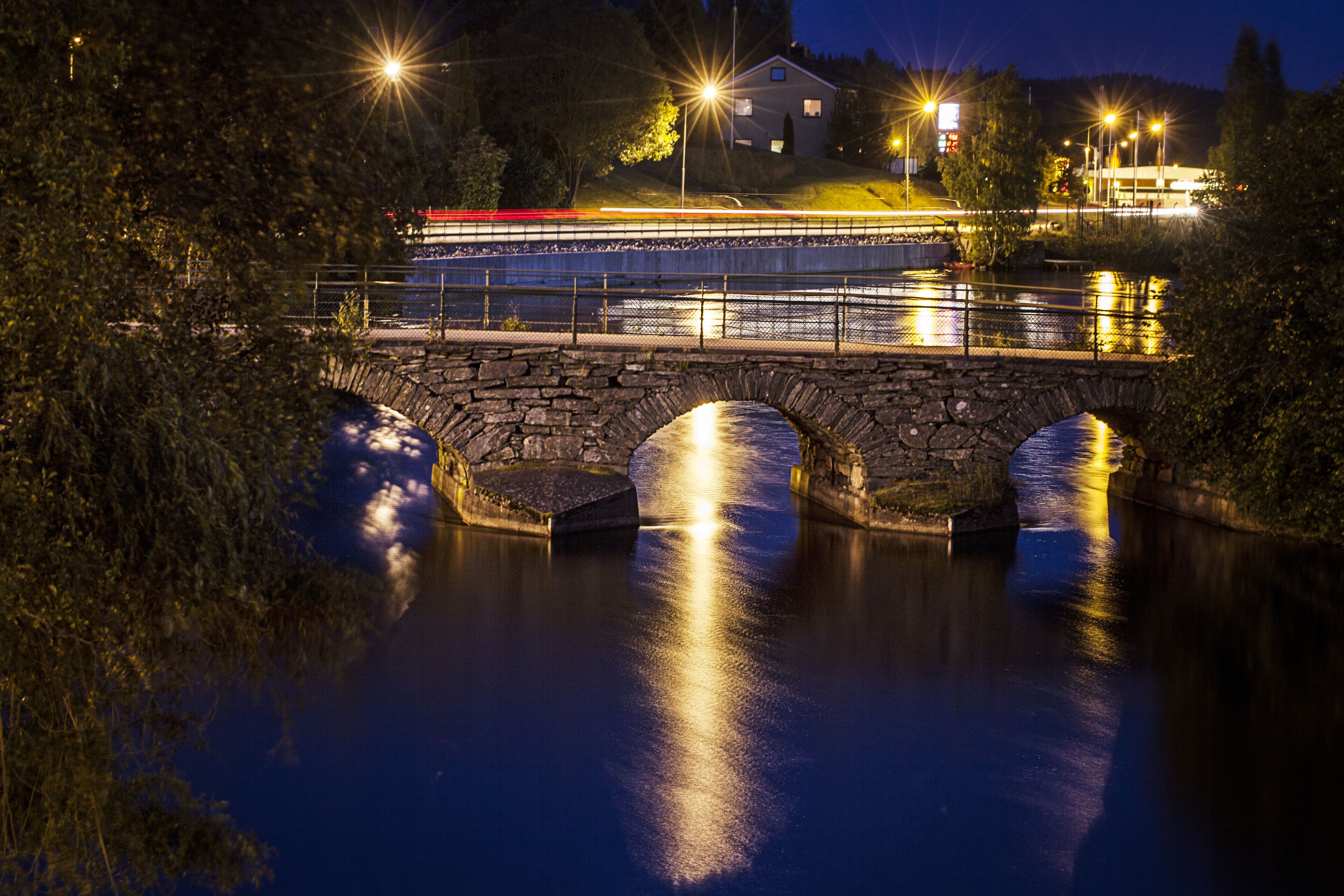 A stone bridge over a calm river at night with streetlights reflecting on the water.