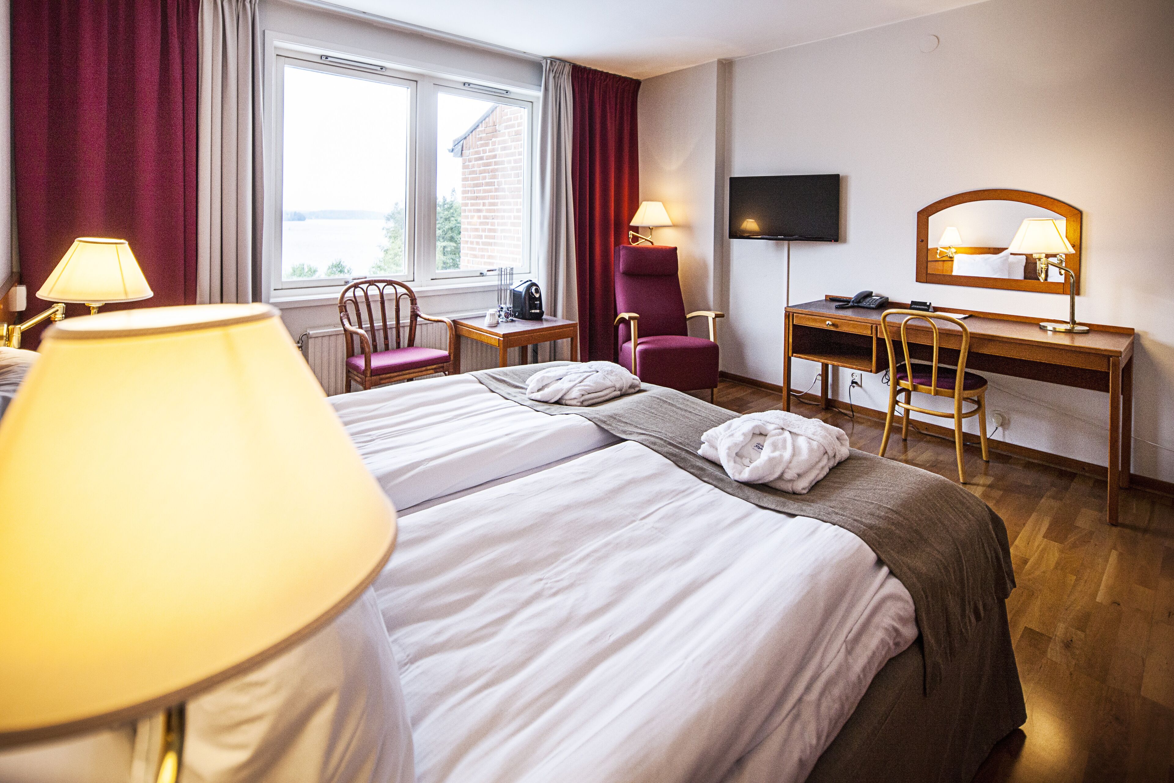 Ein Hotelzimmer mit einem Bett, zwei Stühlen, einem Schreibtisch, einem Fernseher und einem Fenster mit Vorhängen.