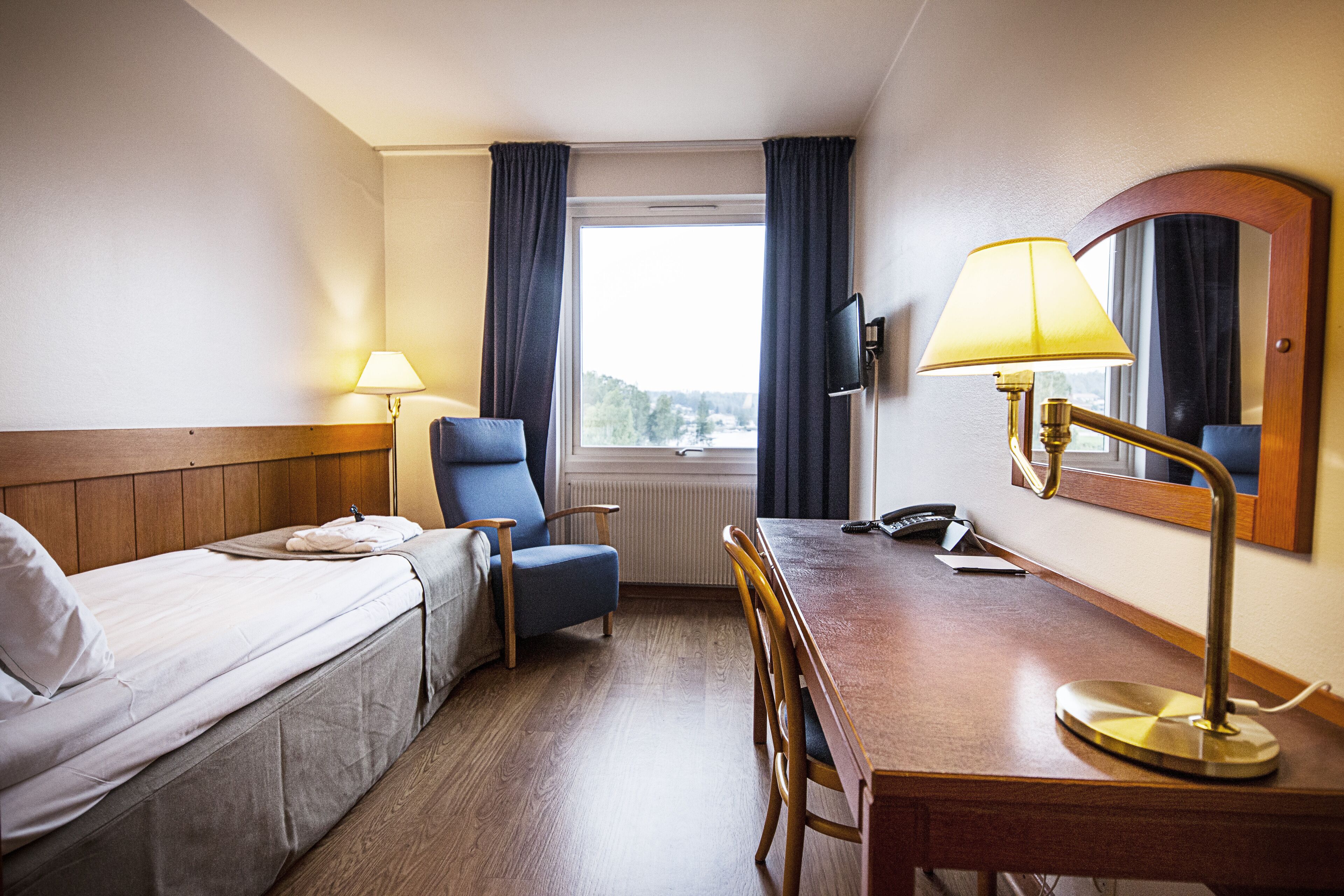 Habitación de hotel con cama, escritorio, silla, lámpara, televisor y ventana con cortinas.