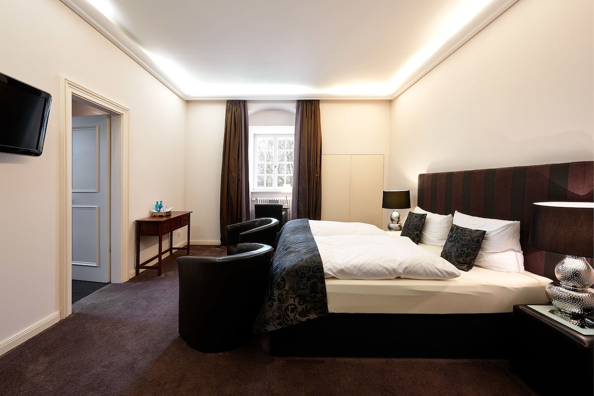Habitación de hotel con cama, escritorio, silla, televisión y ventana con cortinas.