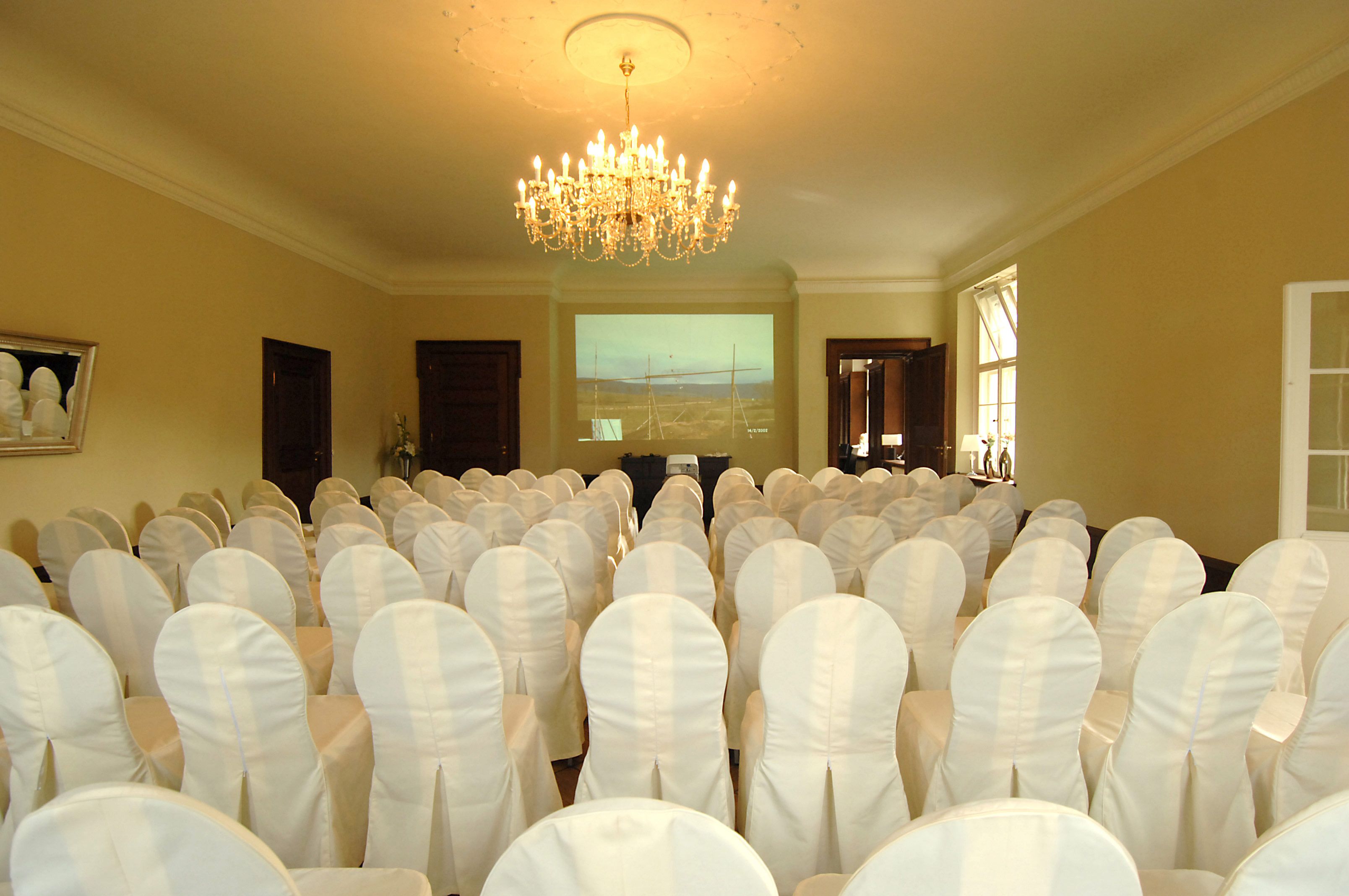 Un salón de conferencias con hileras de sillas blancas, un candelabro y una pantalla de proyección.
