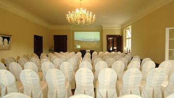 Un salón de conferencias con hileras de sillas blancas, un candelabro y una pantalla de proyección.