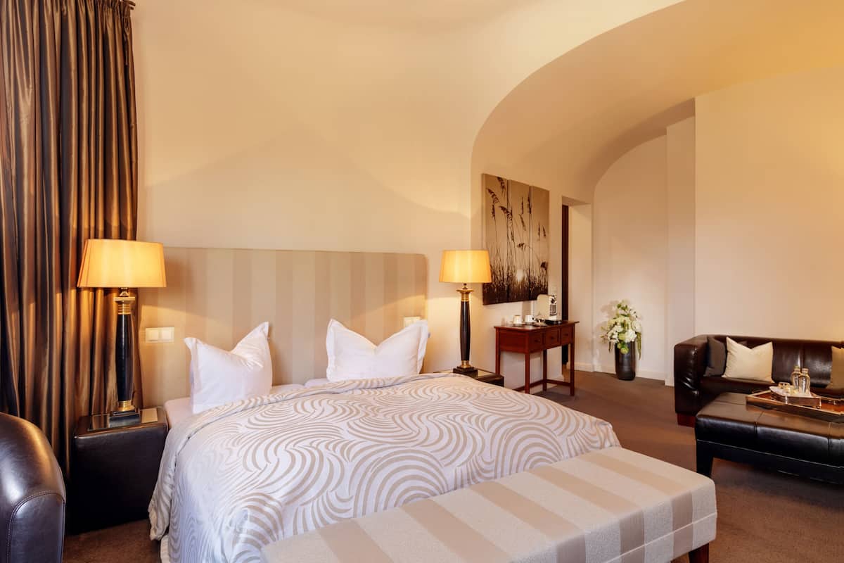 Habitación de hotel con una cama grande, dos mesitas de noche con lámparas, un sofá, una pequeña mesa con un florero y un cuadro en la pared.