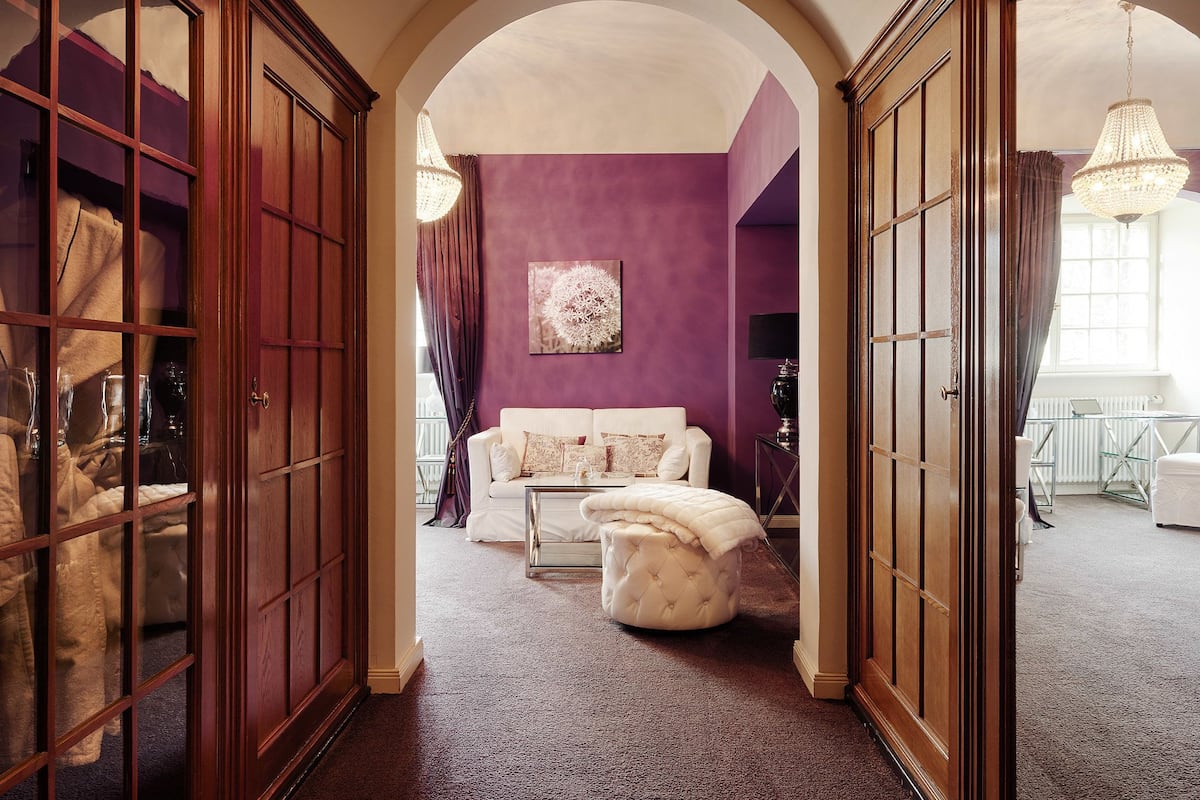 Una habitación con una pared morada, un sofá blanco, un reposapiés capitonado y un armario de madera.