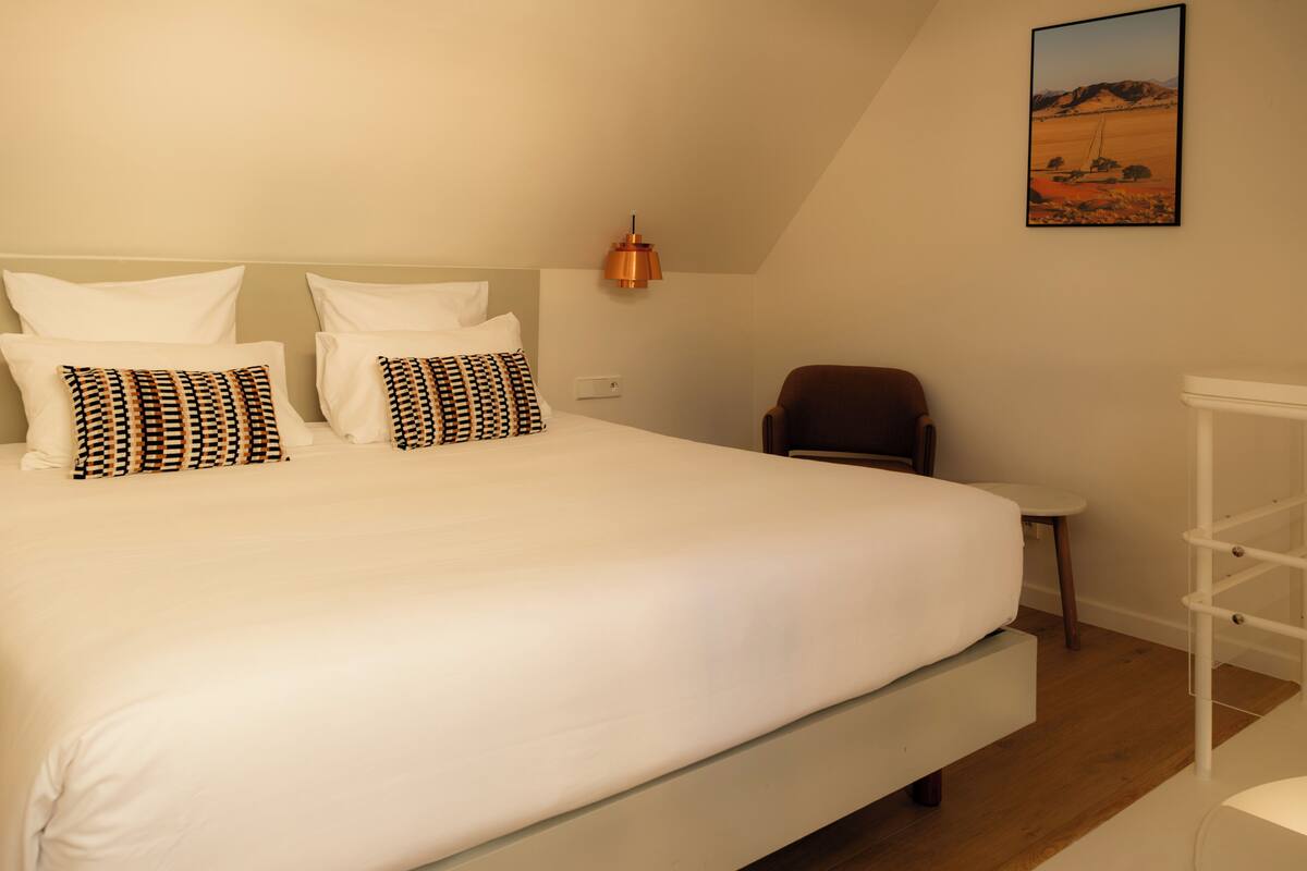 A bedroom with a large bed, a chair, a small table, and a wall-mounted shelf.