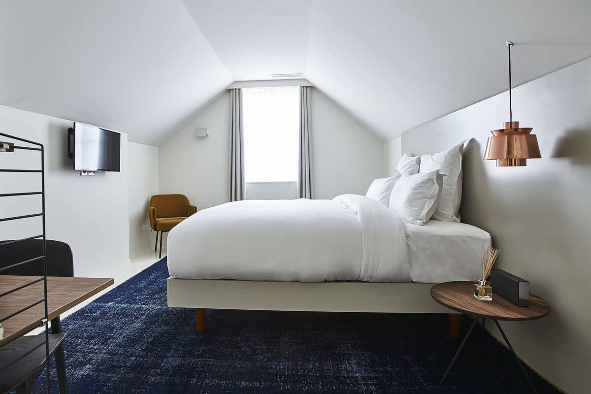 A modern hotel room with a bed, a TV, a chair, a small table, and a lamp.