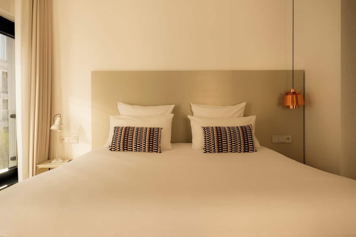 A hotel room with a large bed, two patterned pillows, a bedside lamp, and a hanging pendant light.