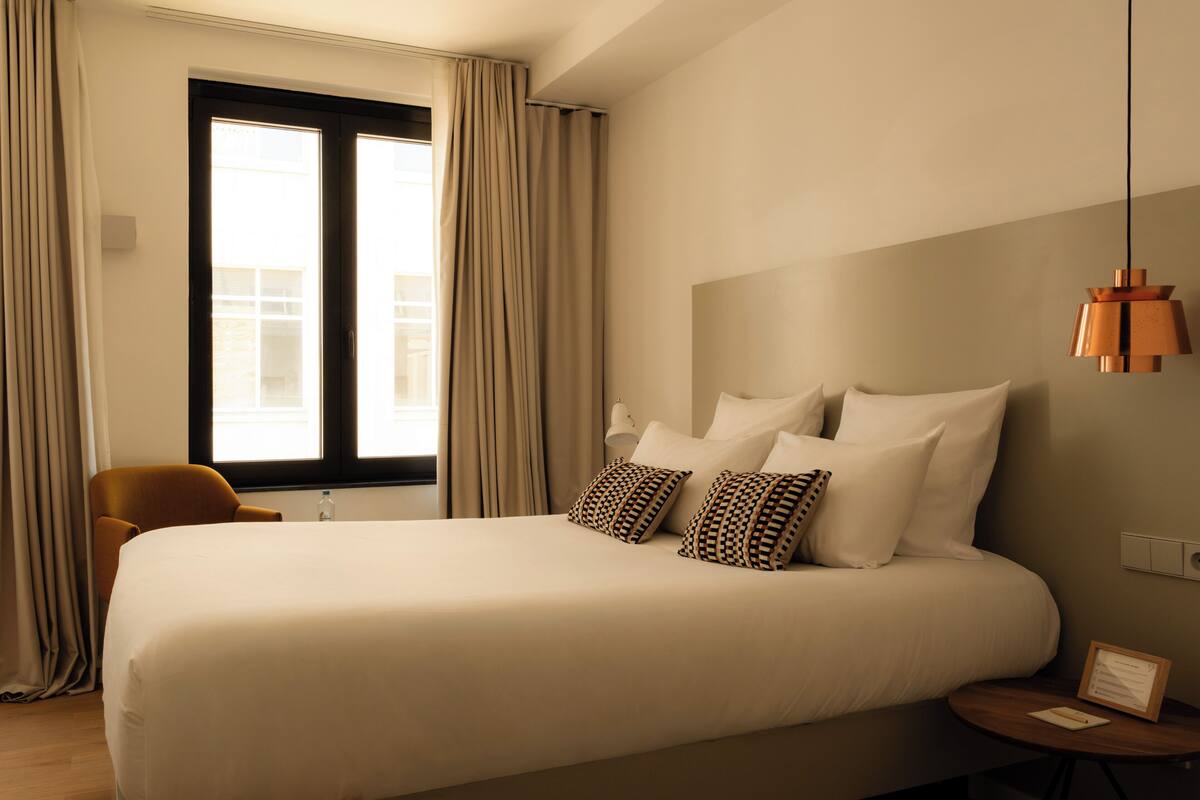 A hotel room with a large bed, a chair, a bedside table, a lamp, and a window with curtains.