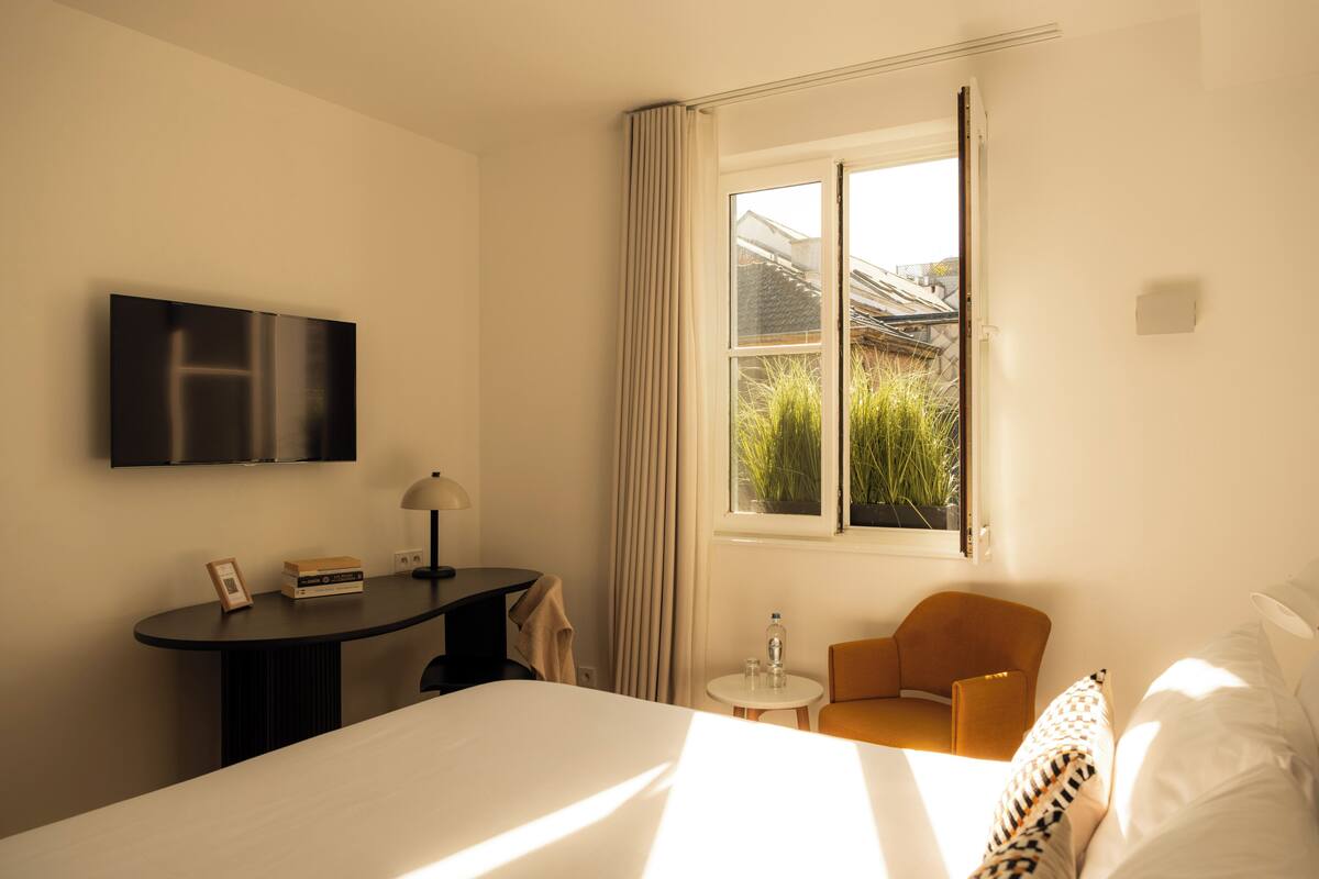 A hotel room with a bed, a desk, a chair, a TV, and a window with a view of greenery.