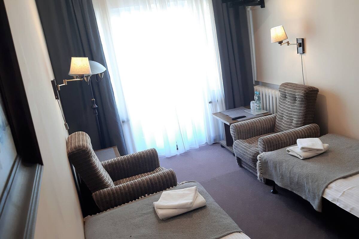 A hotel room with a bed, two armchairs, a table, a lamp, and a view of the outdoors through a window with curtains.