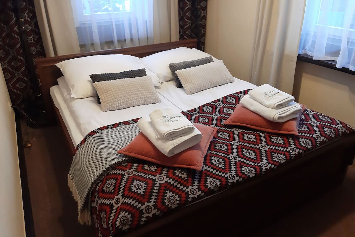 A neatly made bed with a patterned blanket and pillows, a bedside table with a lamp, and a window with sheer curtains.