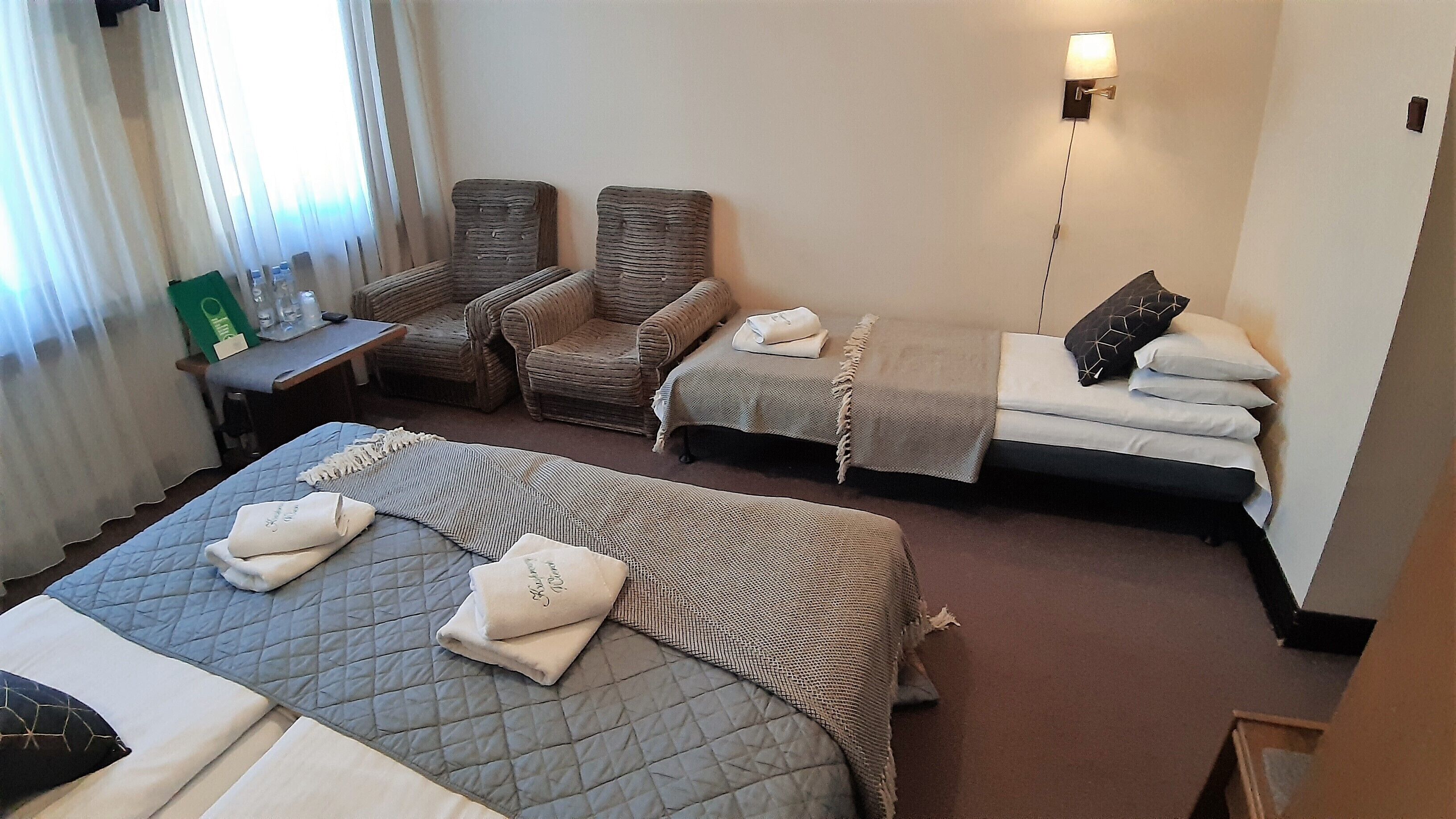 A hotel room with a large bed, two smaller beds, a chair, a table with toiletries, and a wall-mounted lamp.