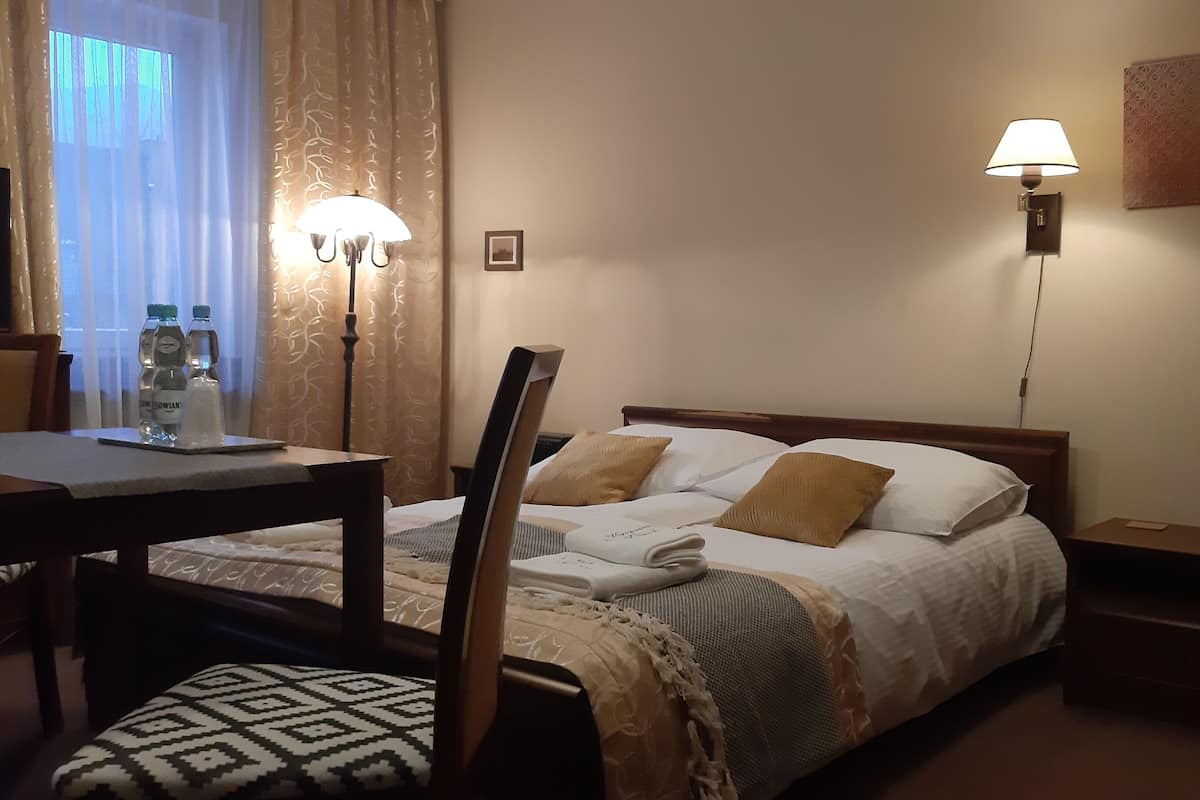 A hotel room with a bed, bedside tables, a desk, and a chair. There are two wall-mounted lamps and a window with curtains.