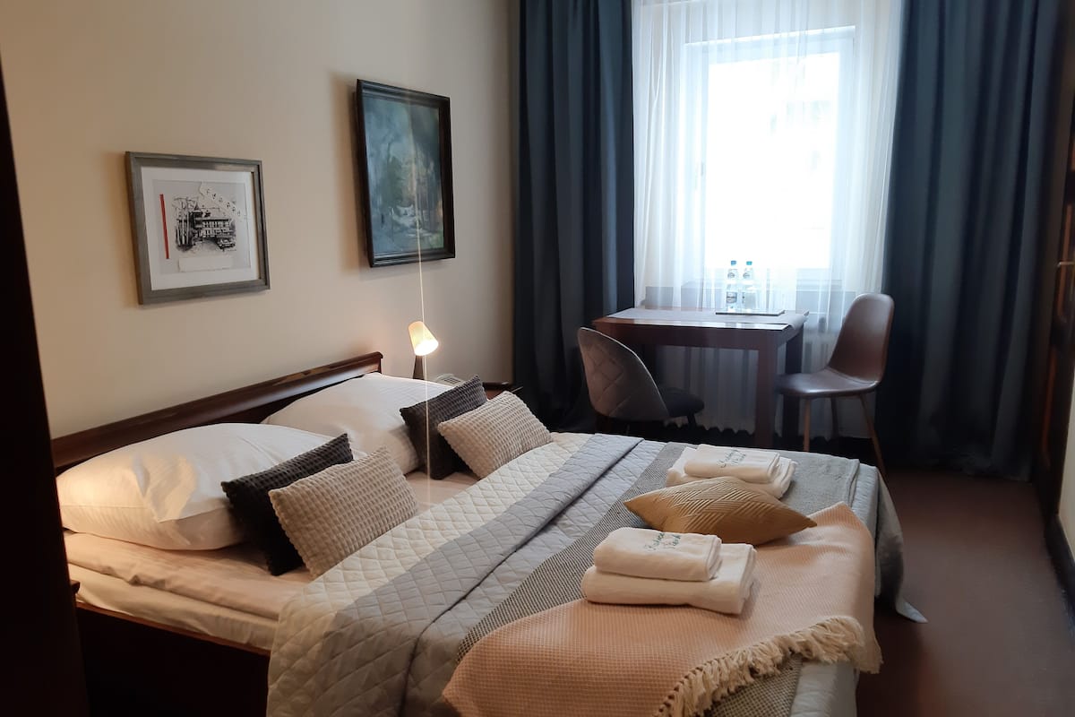 A hotel room with a bed, a desk, a chair, and two framed pictures on the wall.