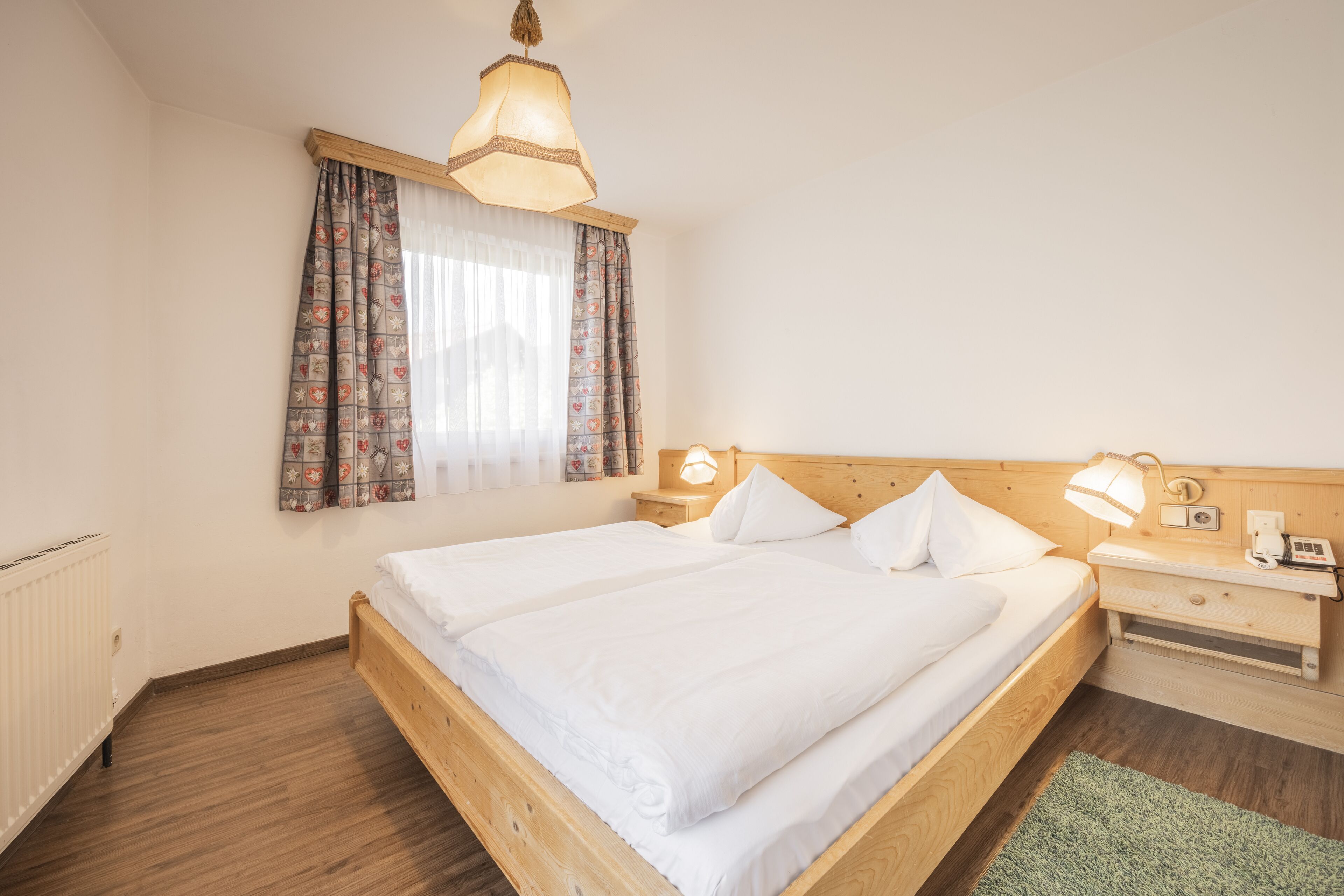 Una cama bien hecha con sábanas blancas, cabecera de madera, mesita de noche con lámpara y una ventana con cortinas estampadas.