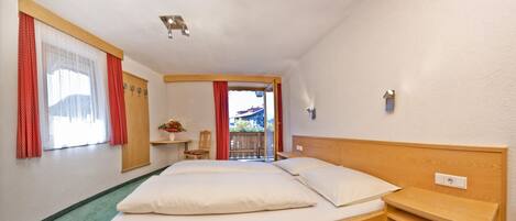 A hotel room with a wooden headboard, a bed with white linens, a small wooden nightstand, a chair, and a window with red curtains.
