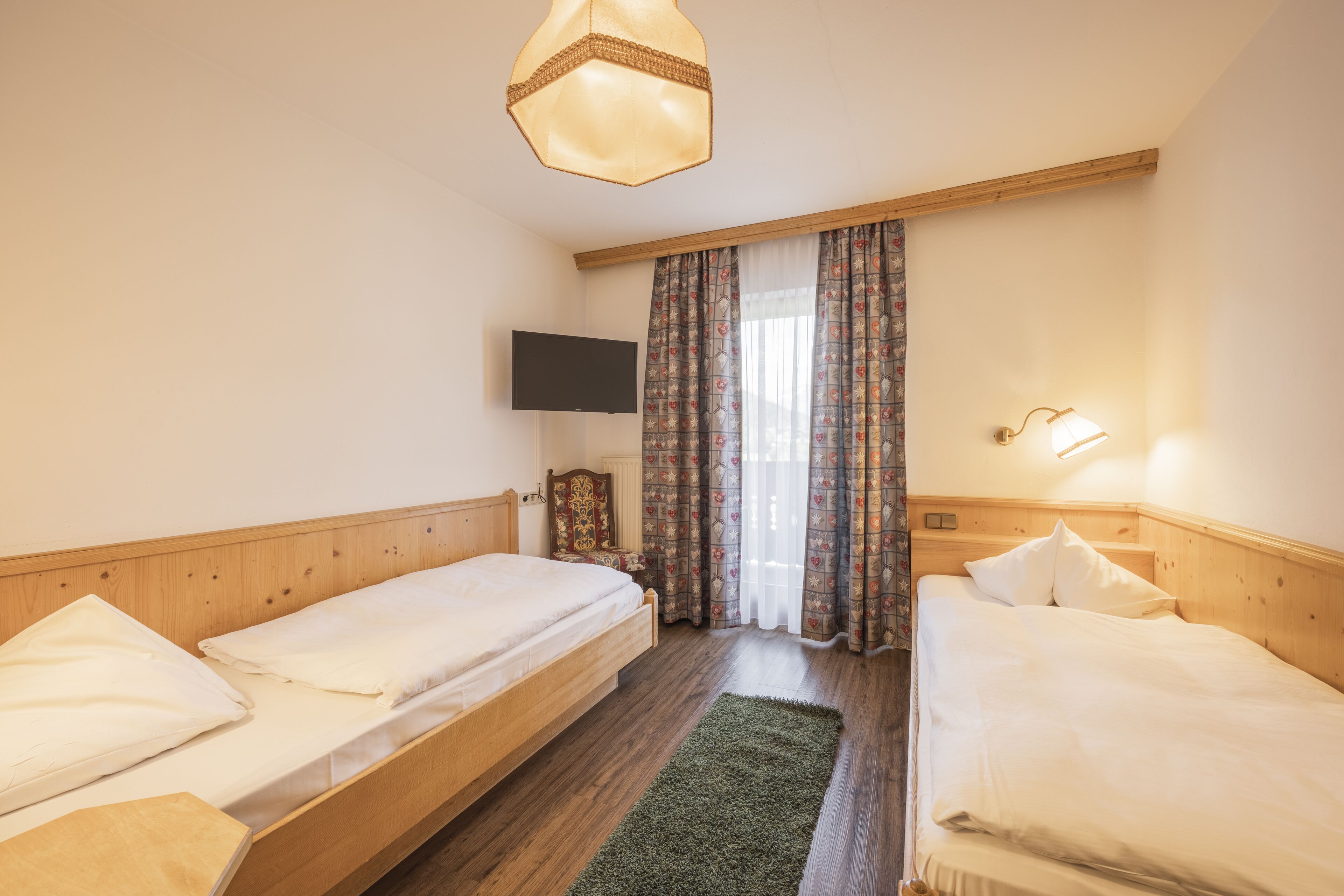 Ein Schlafzimmer mit zwei Einzelbetten, einem an der Wand befestigten Fernseher, einem Holz-Kopfteil und einem Fenster mit Vorhängen.