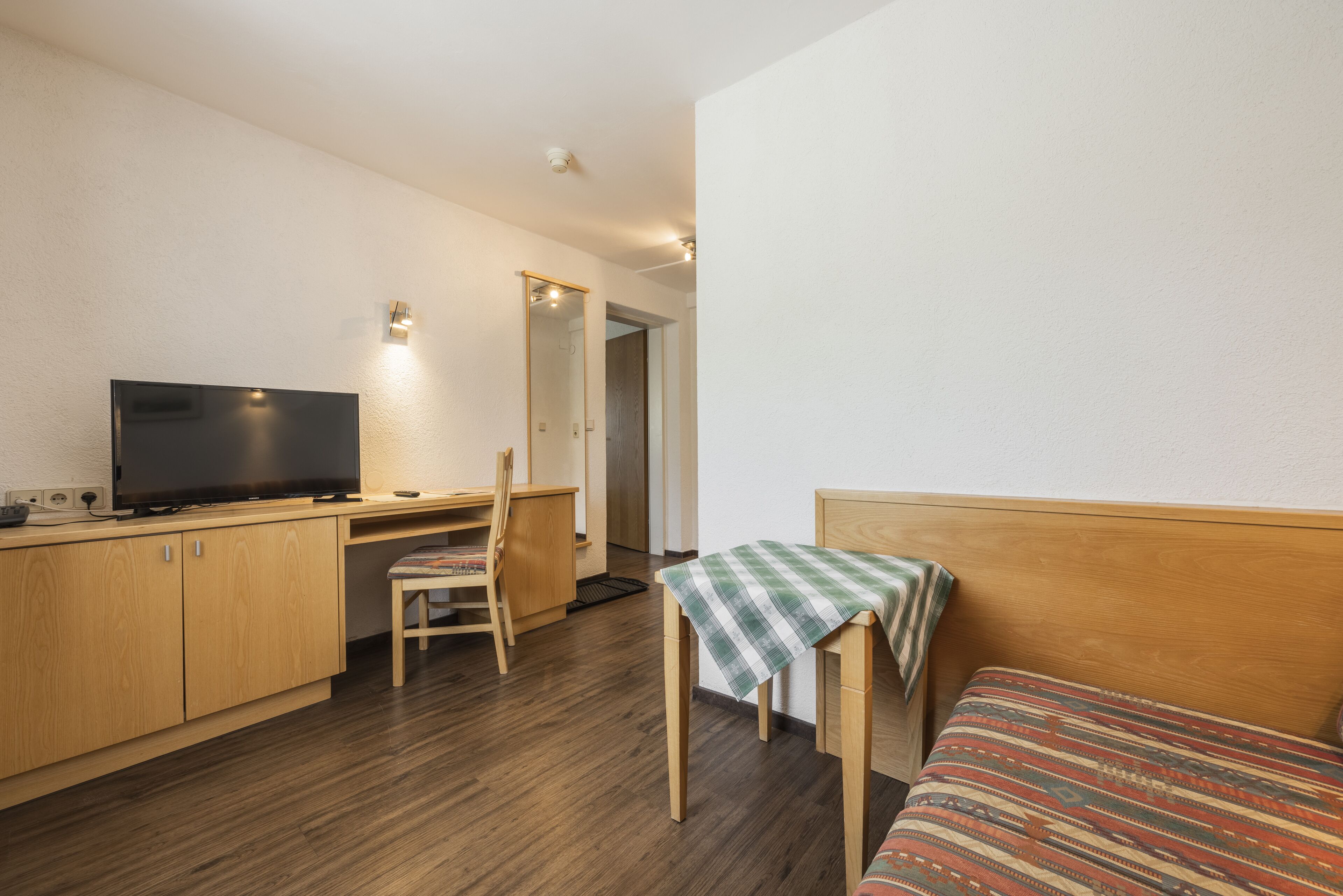 Une pièce de taille modeste, comprenant un bureau en bois, une télévision, une chaise, un lit et une petite table recouverte d’une nappe.