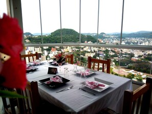 A dining table set for two with a city view, a flower vase, and a laptop on the table. - Hotel Farol (Joacaba)