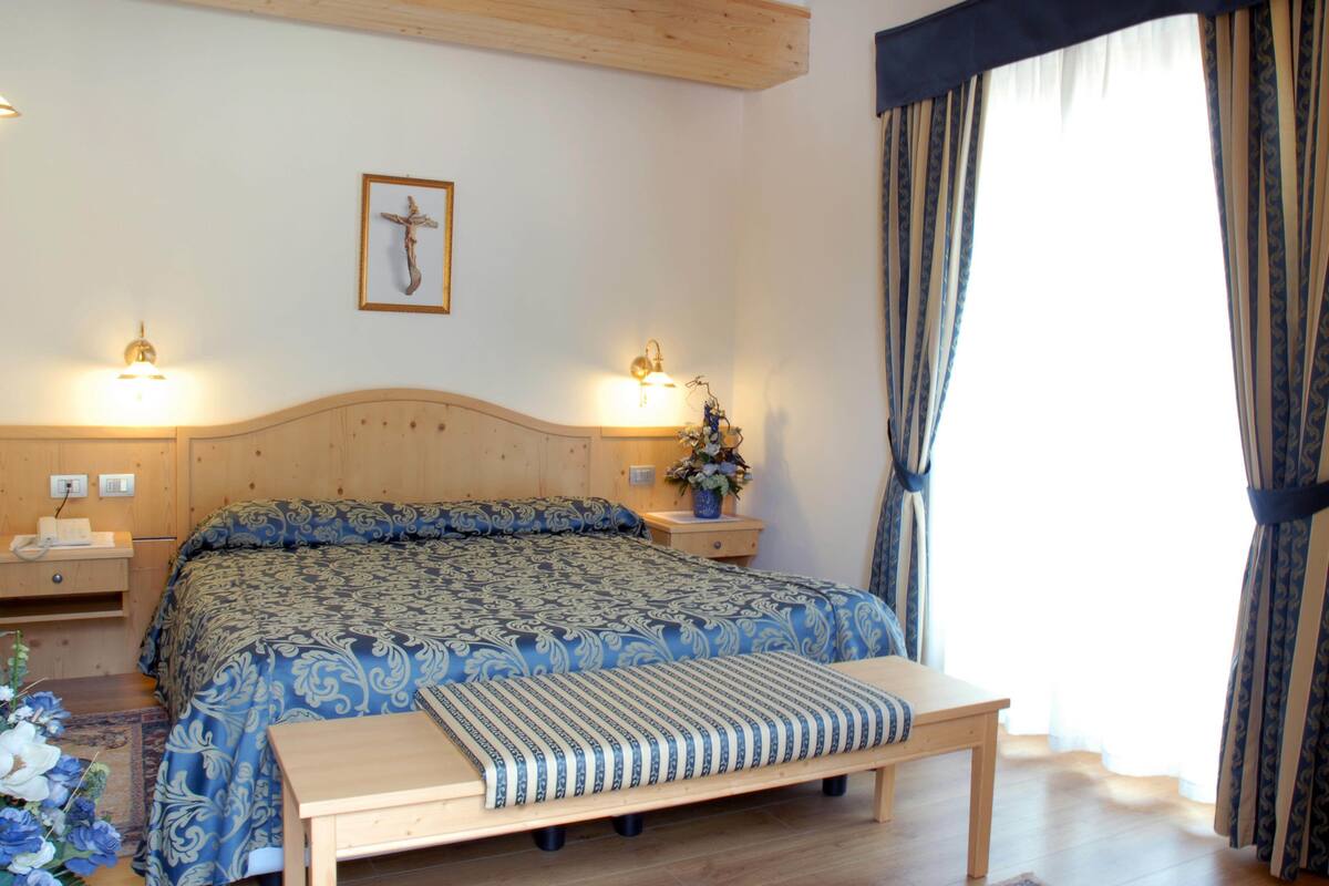 Een slaapkamer met een houten hoofdbord, een groot bed met beddengoed met een patroon, bedlampjes, een nachtkastje met een vaas bloemen en een raam met gordijnen.