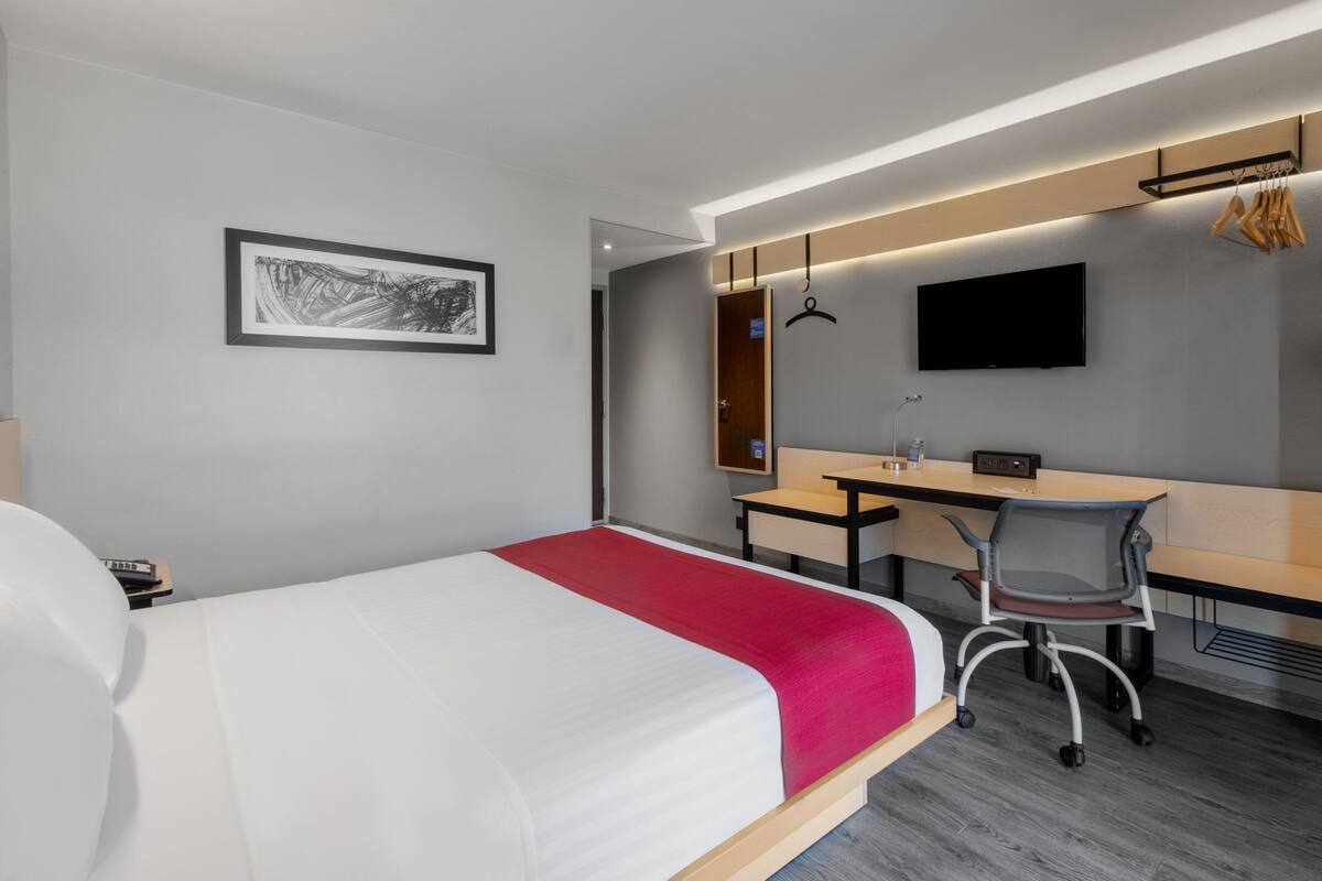 Ett modernt hotellrum med en stor säng, ett skrivbord, en stol, en TV och en inramad tavla på väggen.