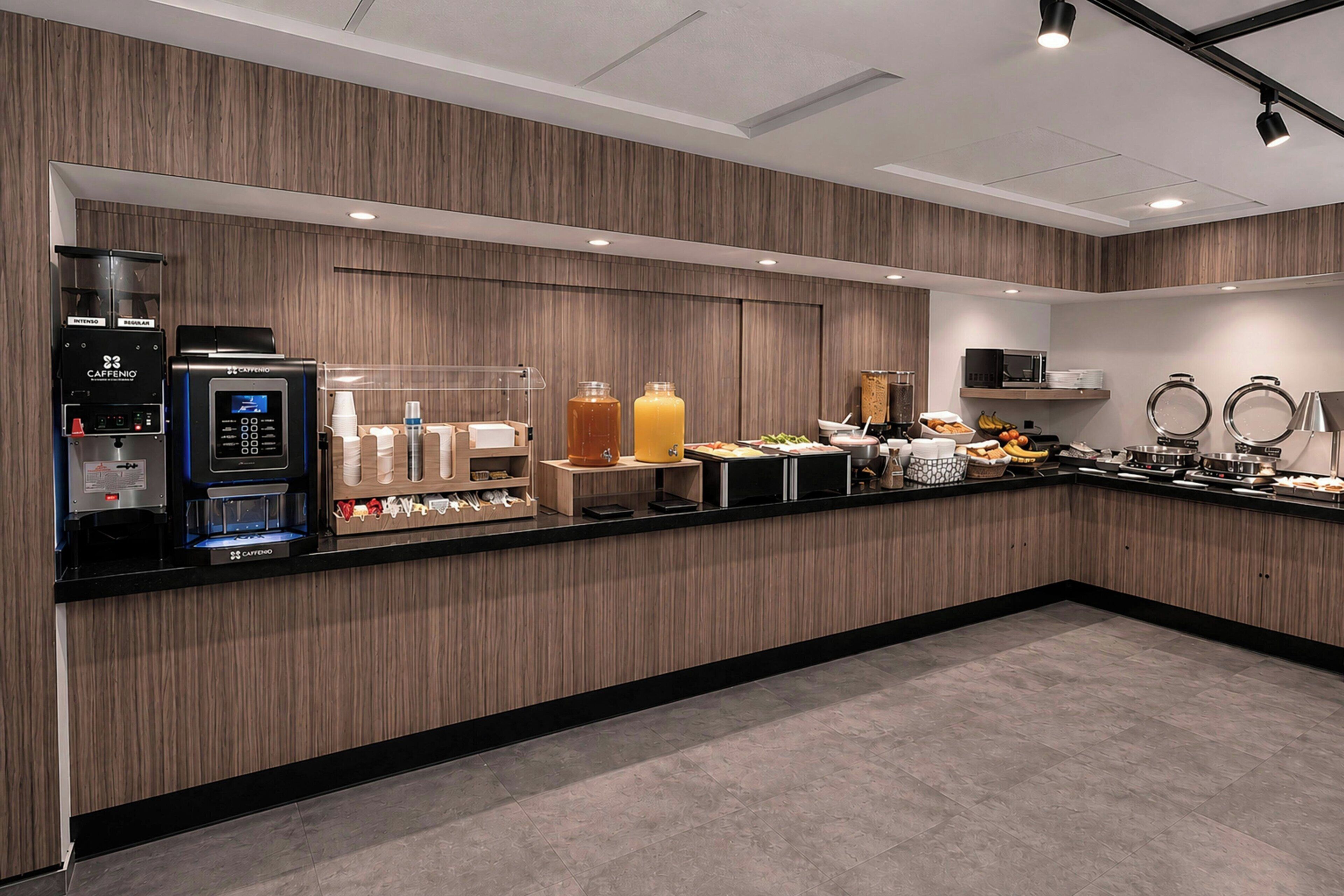 Ein moderner Frühstücksbereich eines Hotels mit Kaffeeautomat, Saftspendern und einer Mikrowelle.