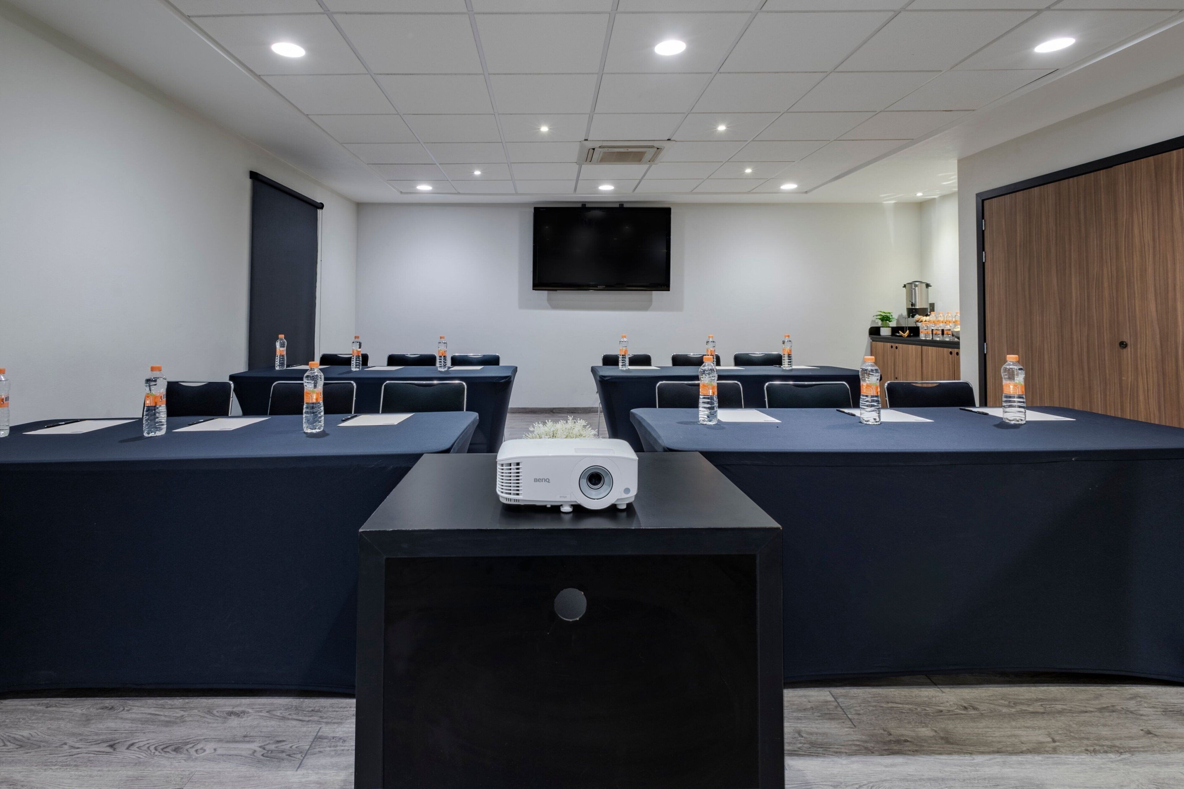 Una sala de conferencias con un proyector, mesas con botellas de agua, un televisor en la pared y un armario de madera.