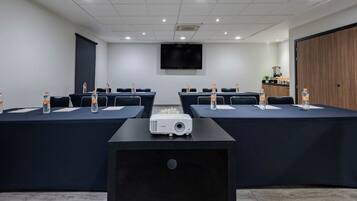 Una sala de conferencias con un proyector, mesas con botellas de agua, un televisor en la pared y un armario de madera.