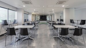 A modern conference room with white tables and black chairs, large windows, and a central black screen.