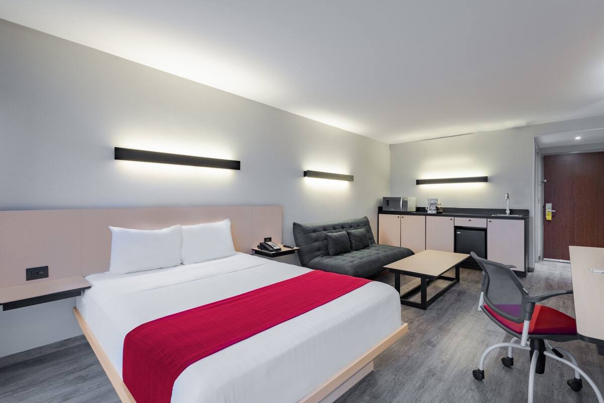 Ett modernt hotellrum med en säng, ett skrivbord, en stol och ett litet kök.