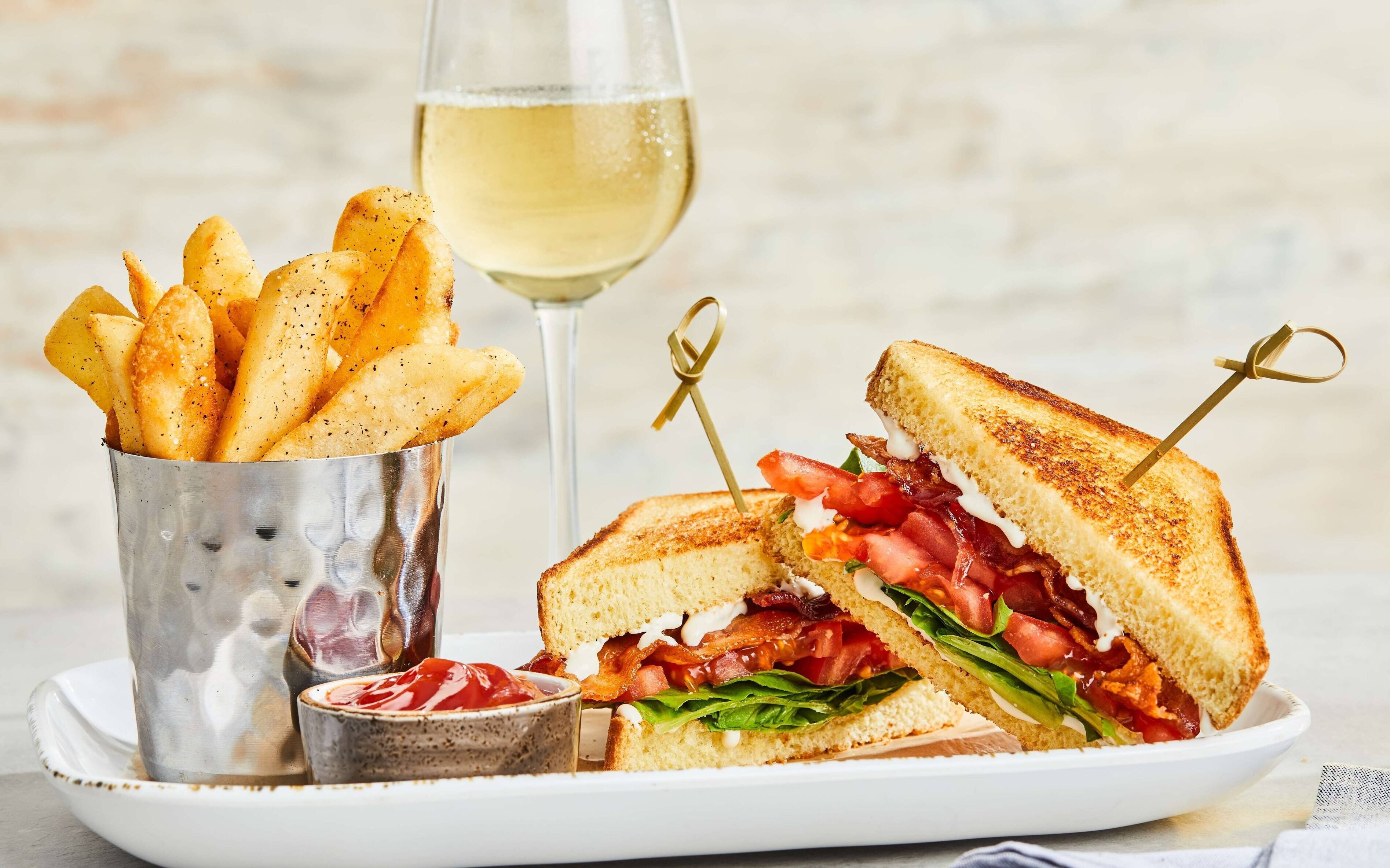 A plate with a grilled sandwich, a glass of white wine, and a container of fries.