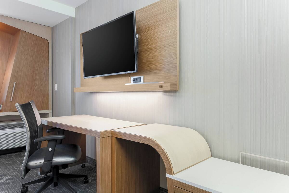 Ein modernes Hotelzimmer mit einem Holztisch, einem schwarzen Bürostuhl, einem flachbildschirmfernseher an der Wand und einer festen Sitzbank.